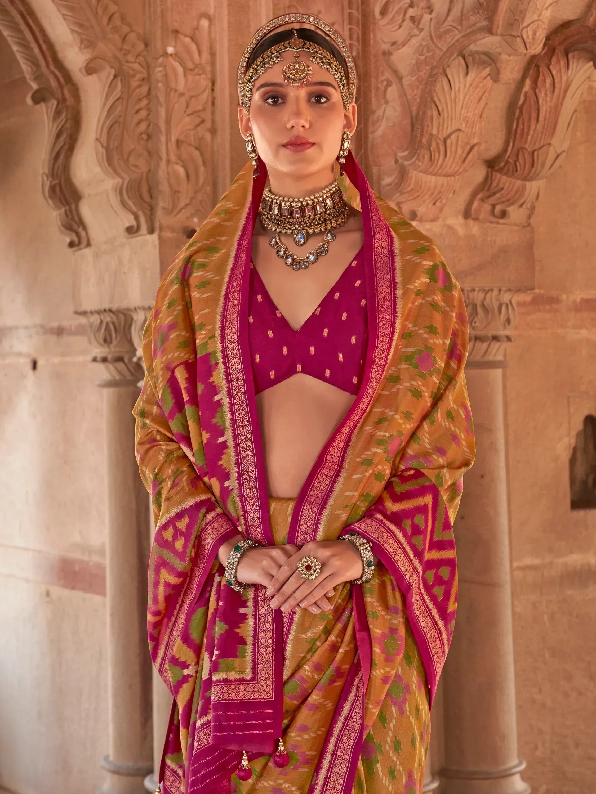 Woman in vibrant pink and gold ethnic saree with traditional jewelry, standing against carved stone architecture
