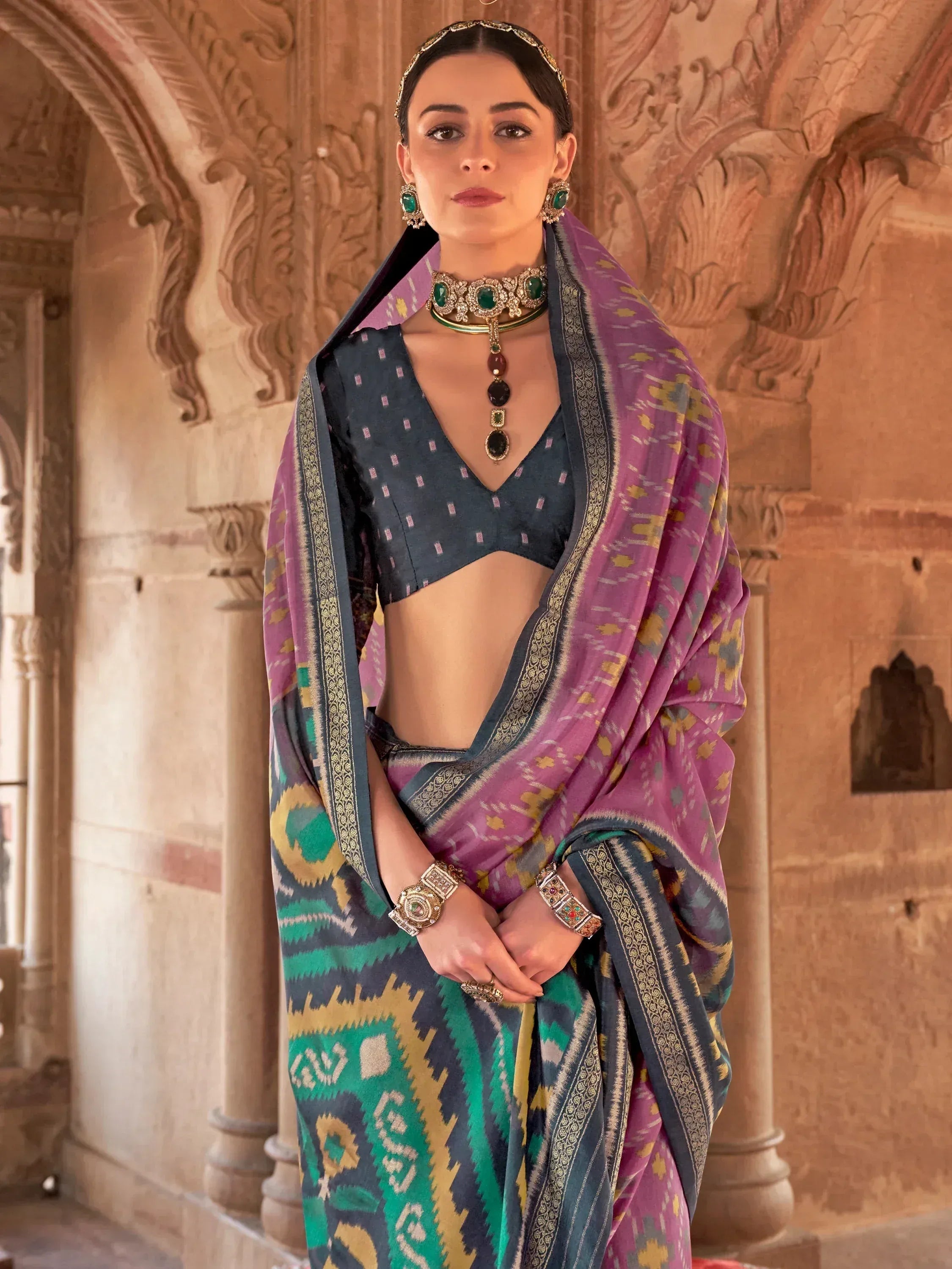 Woman in vibrant purple and green ikat saree with designer blouse, traditional jewelry, against carved stone arches