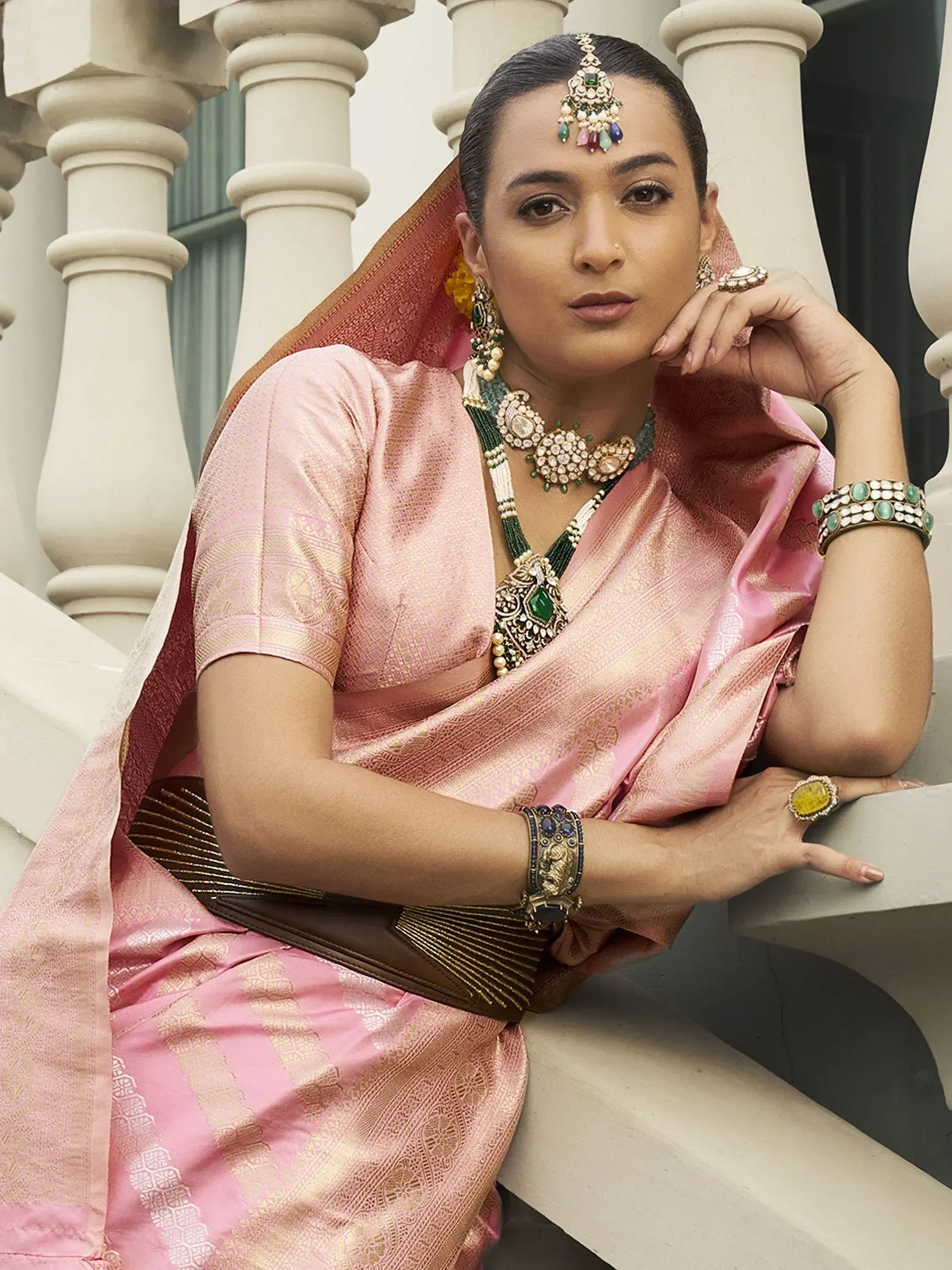 Woman in pink silk saree with gold embroidery, traditional Indian jewelry, and temple-style necklace, posing on white balustrade