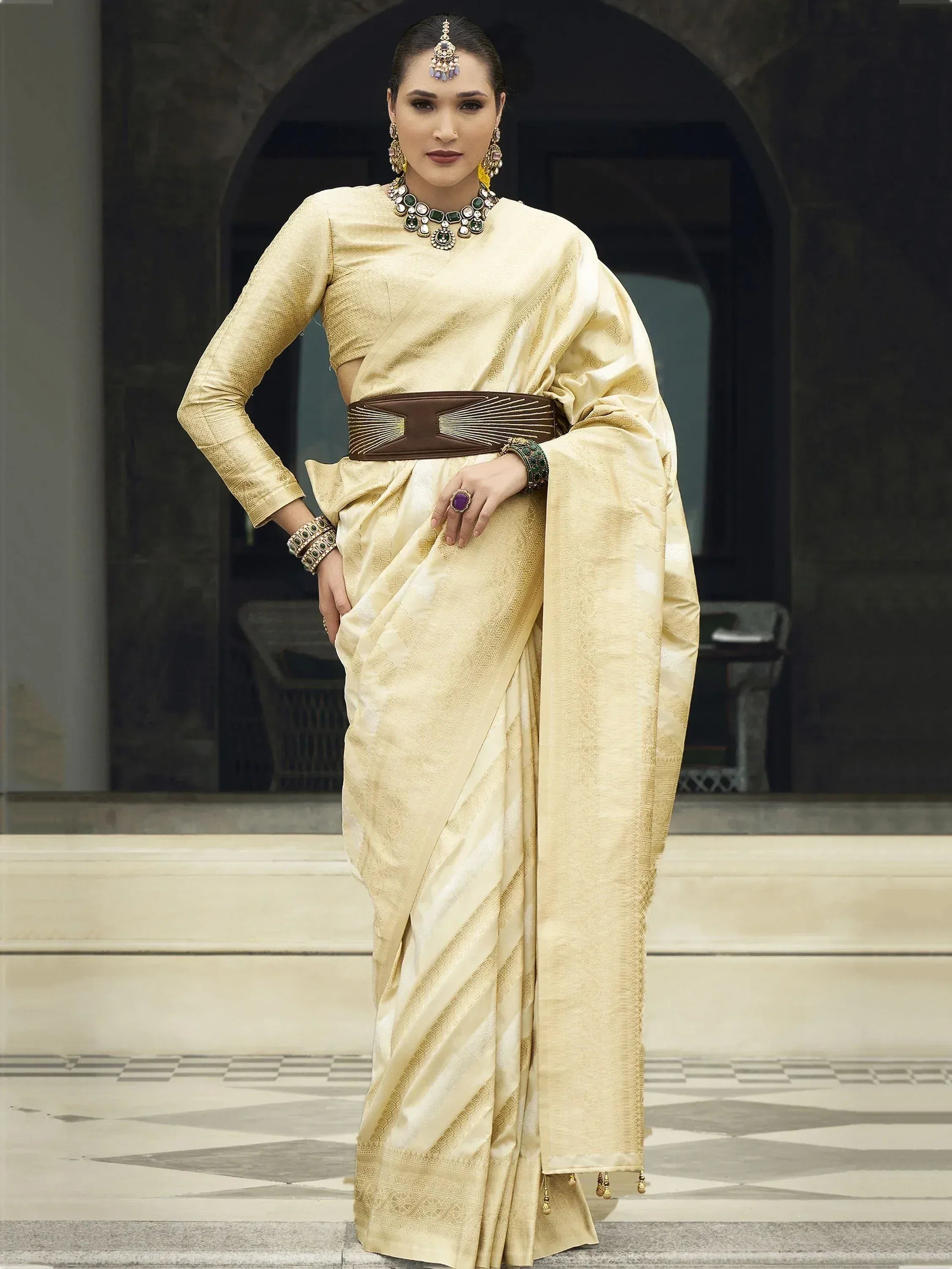 Elegant cream saree with gold weave, paired with statement jewelry and a brown belt, perfect for Indian ethnic occasions.