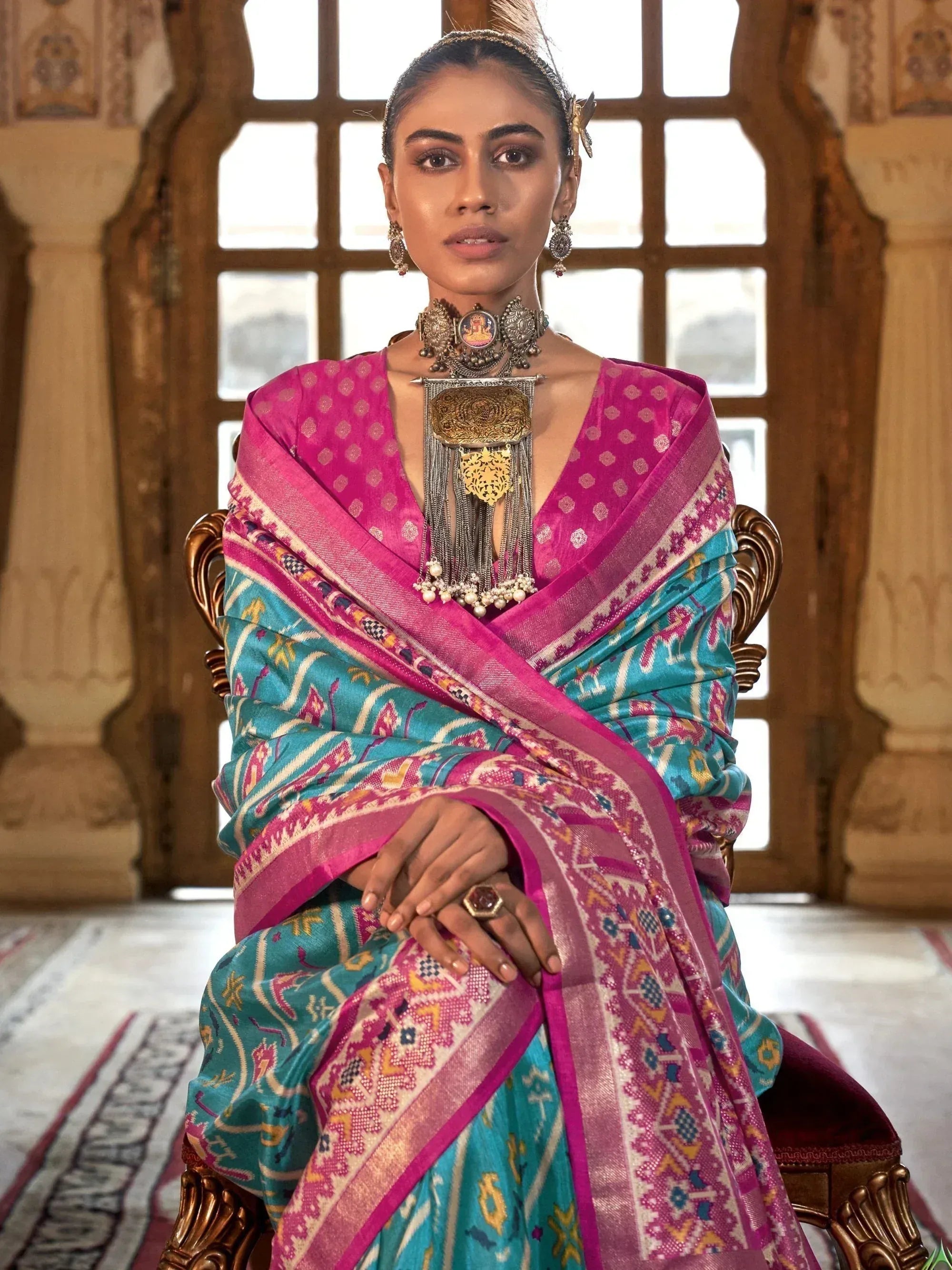 Woman in vibrant pink and teal ethnic saree with intricate patterns, traditional jewelry, seated in ornate palace setting