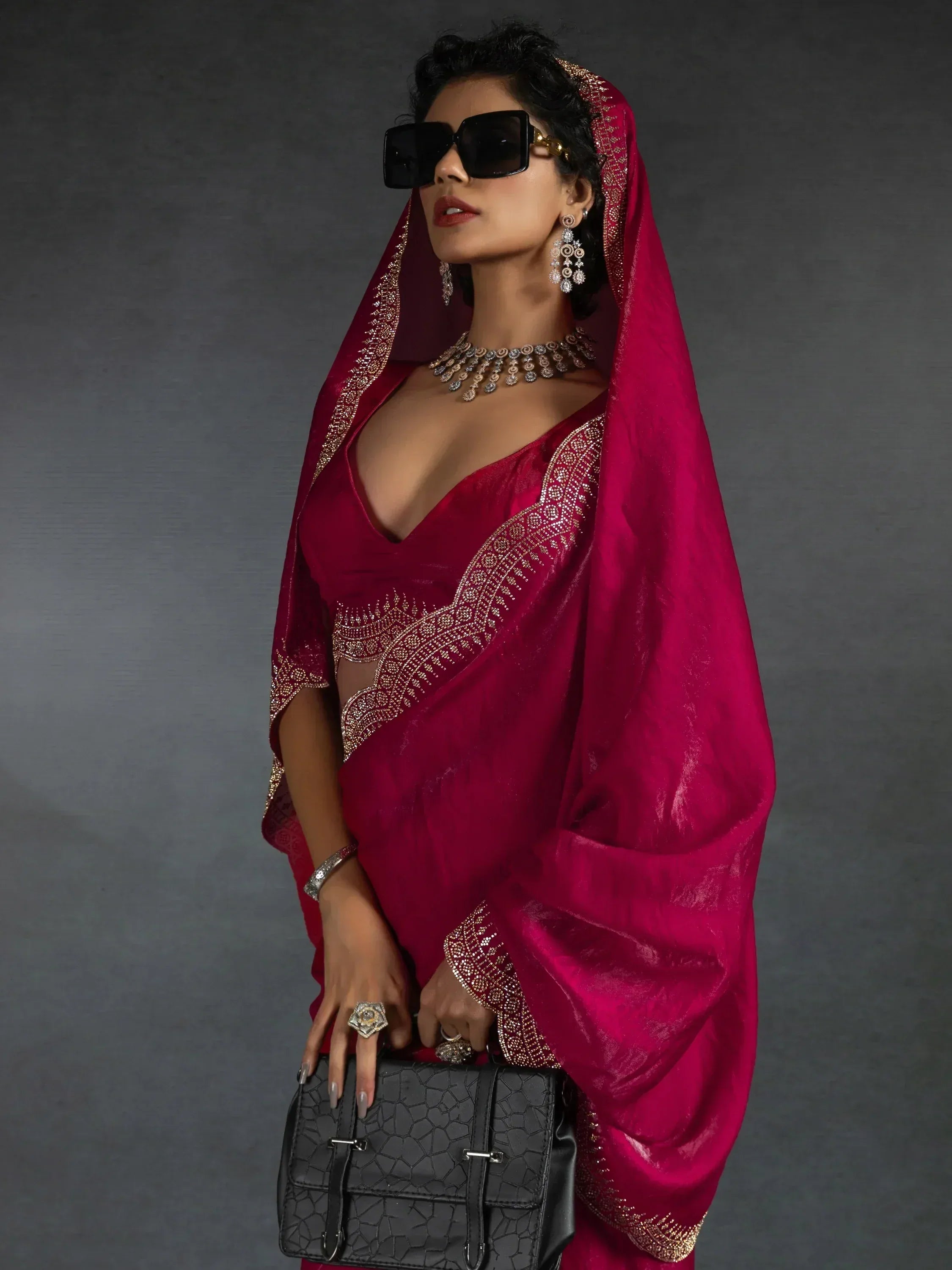 Woman in maroon velvet saree with gold embroidery, statement jewelry, and black sunglasses holding a textured handbag