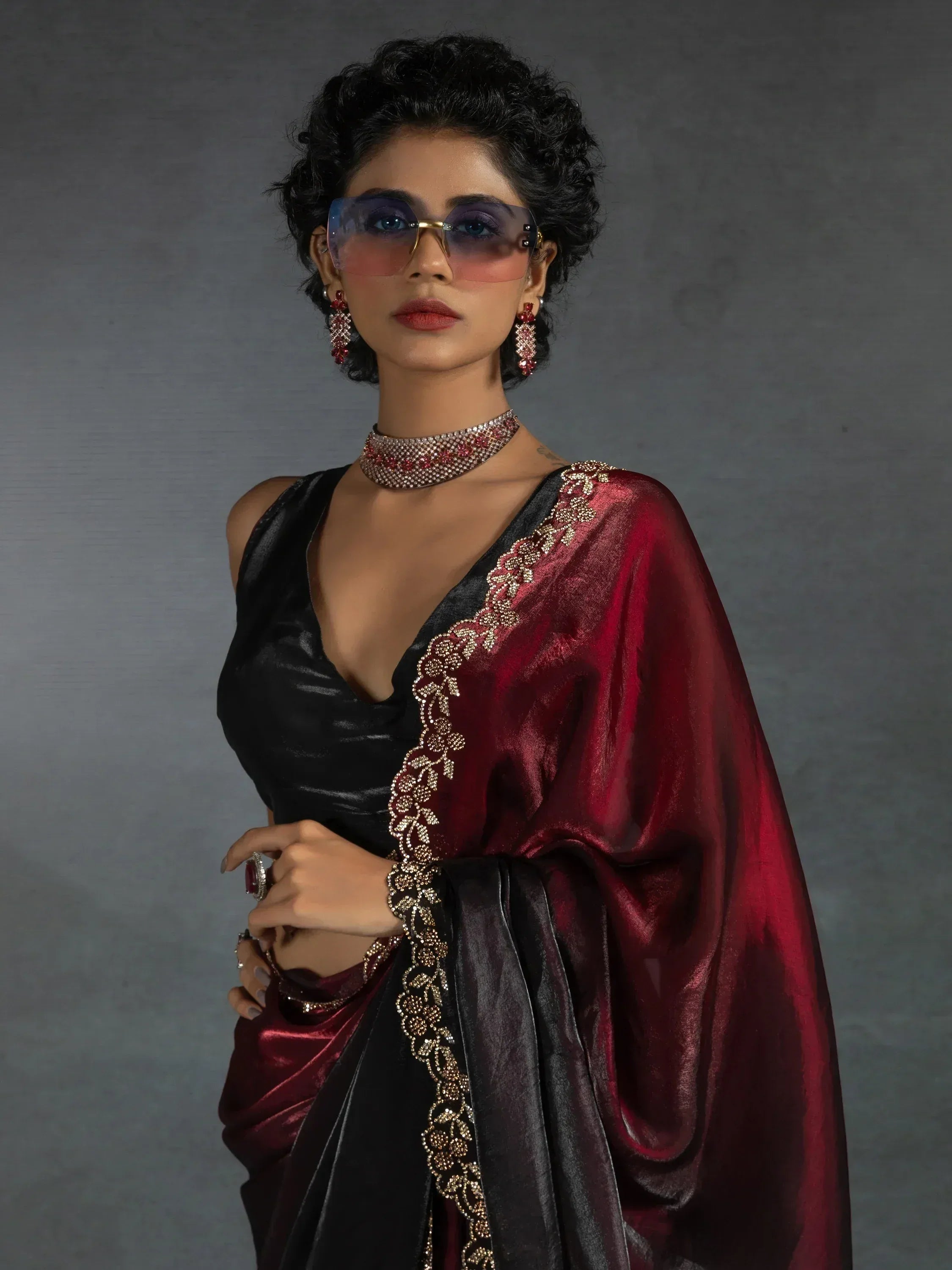 Elegant woman in black and maroon saree with gold embroidery, styled with statement jewelry and sunglasses.