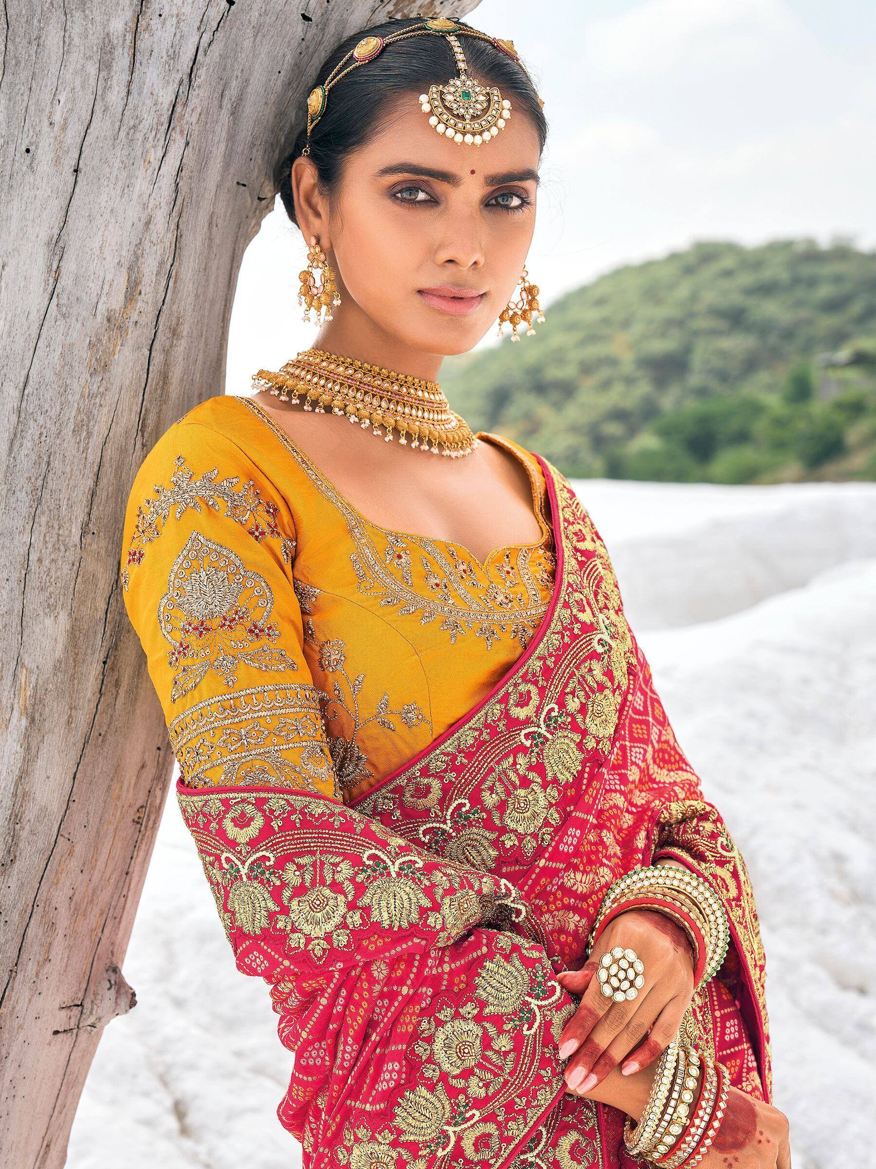 Woman in red and gold embroidered saree with matching jewelry, leaning against tree