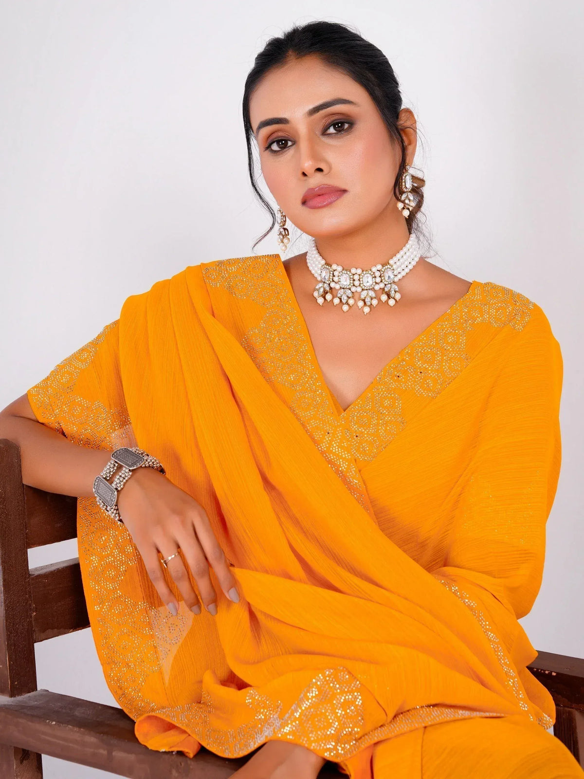 Woman in vibrant yellow ethnic saree with intricate embroidery and pearl jewelry, seated on wooden chair