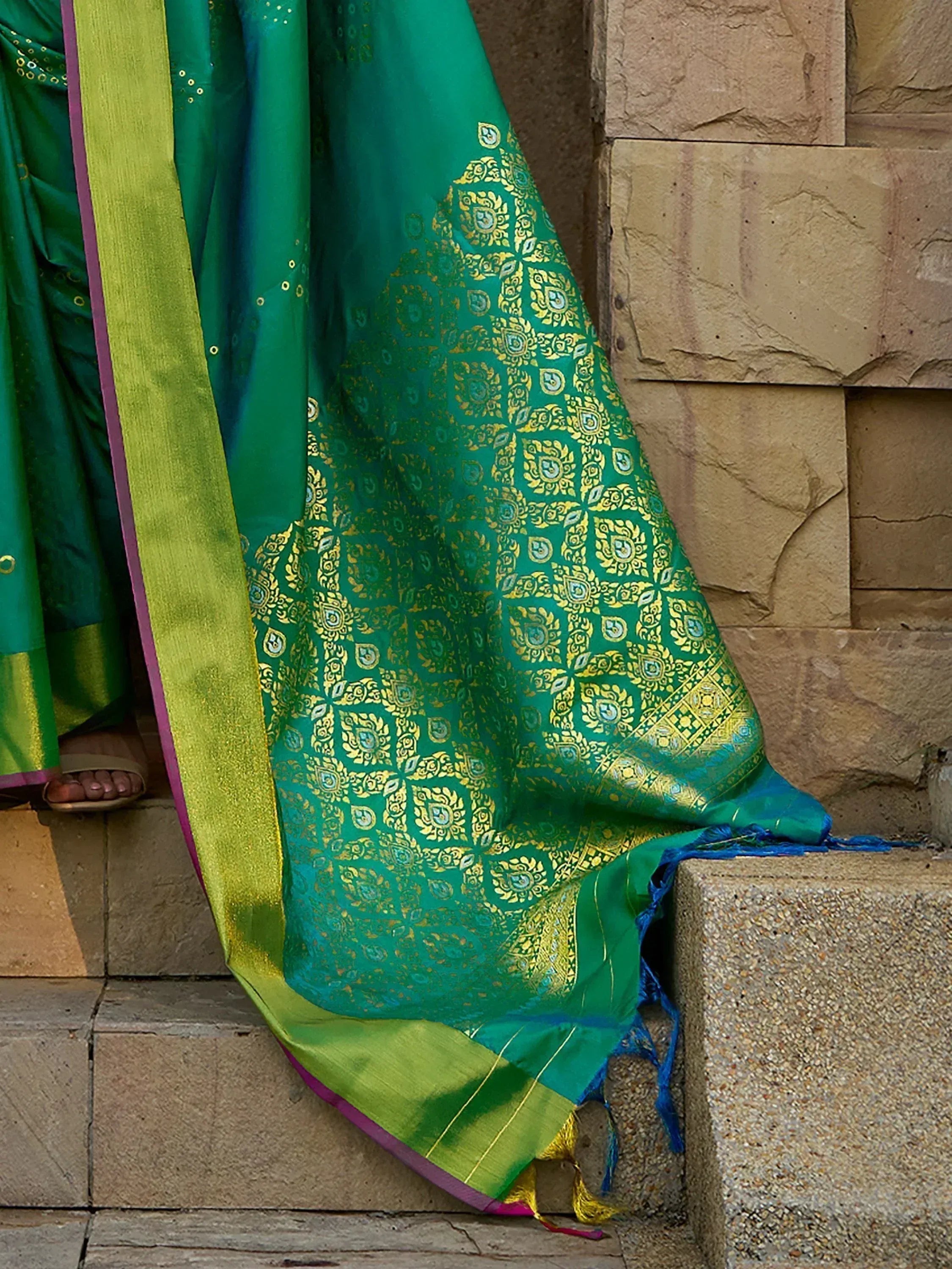 Emerald green saree with gold zari work, lime border, and blue tassels, draped on stone steps