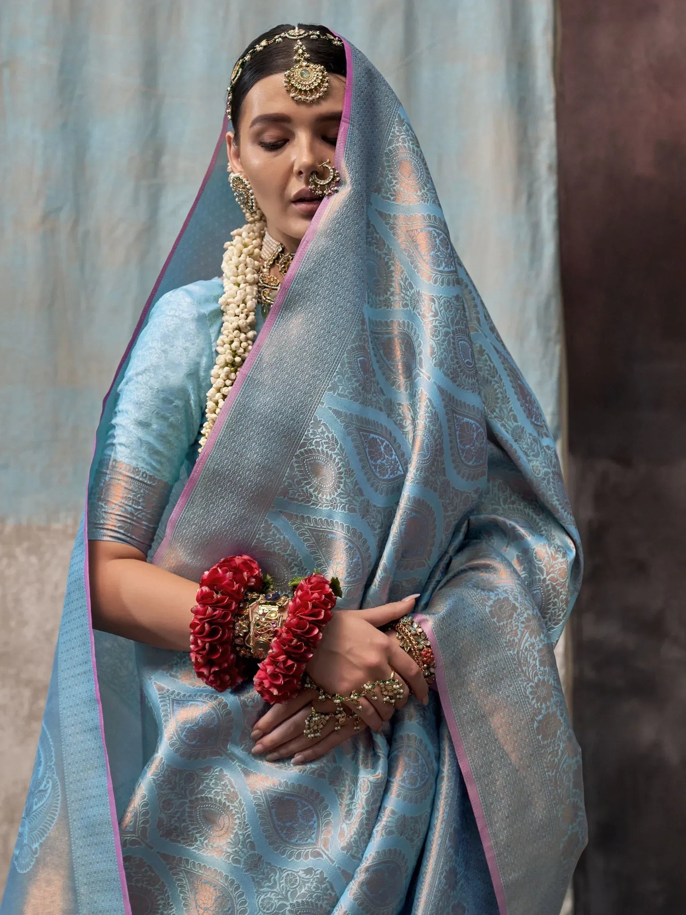 Elegant Indian bride in light blue designer saree with gold paisley print, traditional jewelry, and floral adornments.