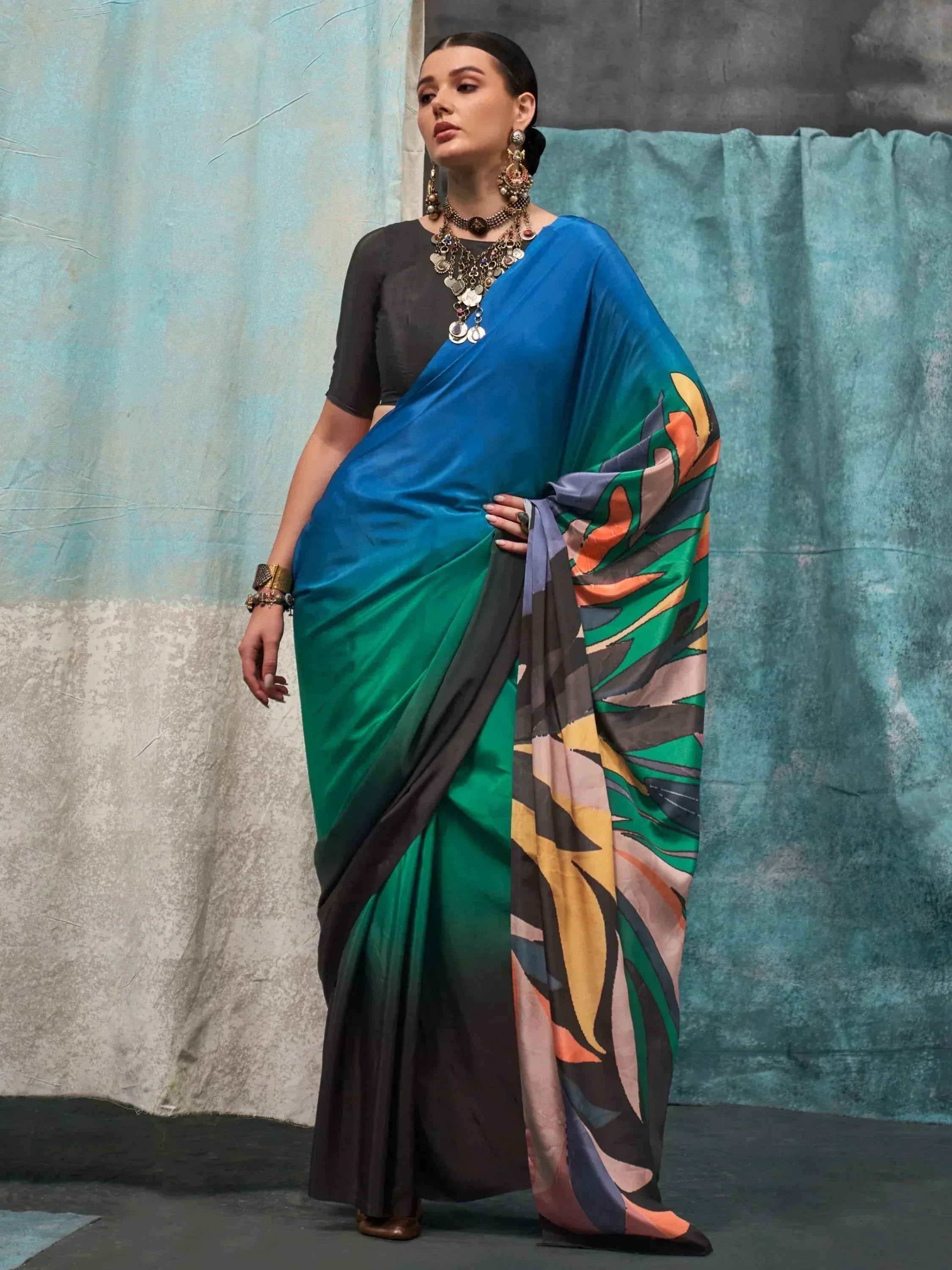 Woman in vibrant blue and green ombre saree with abstract print, styled with black blouse and ethnic jewelry