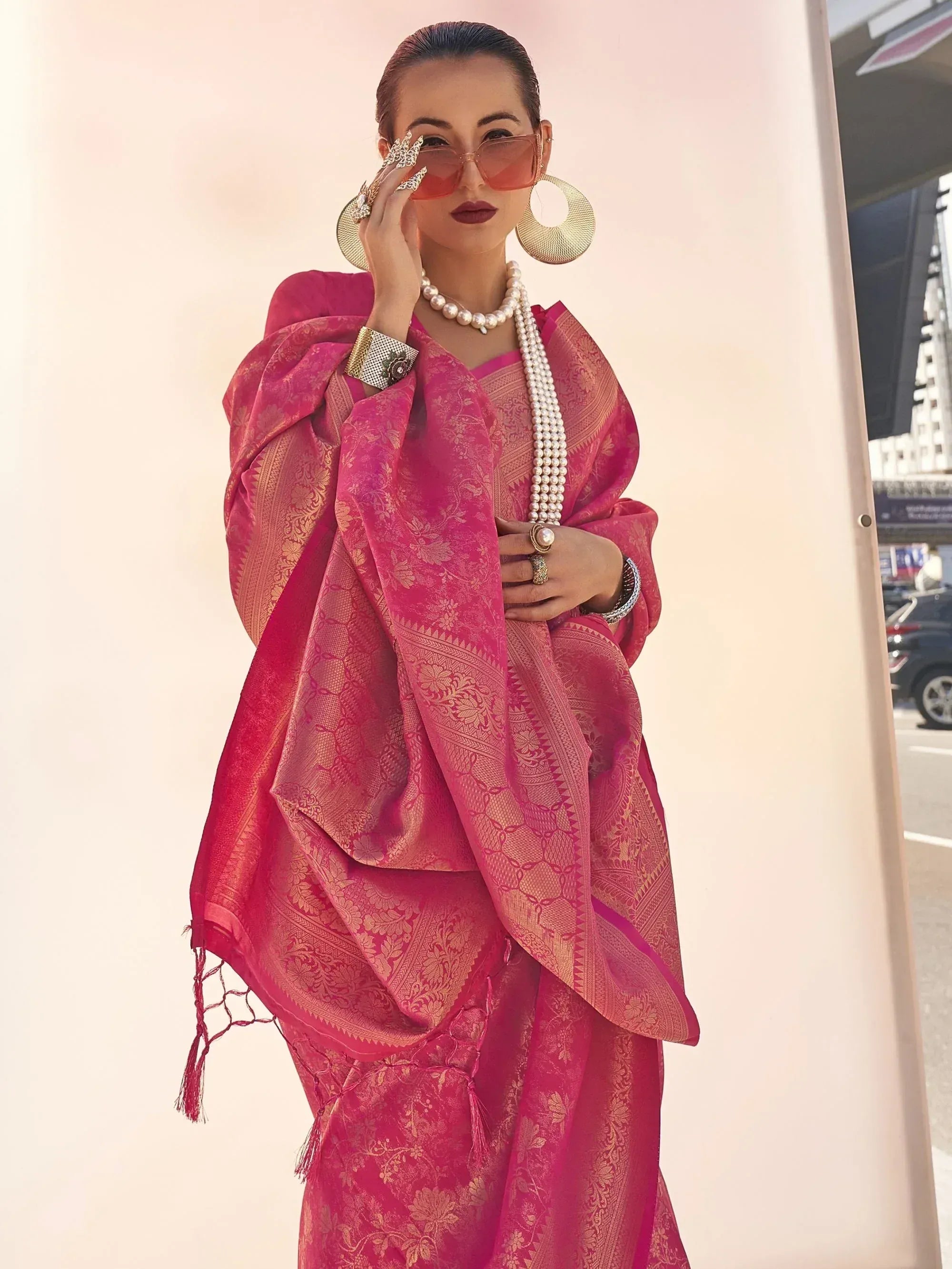 Woman in vibrant pink ethnic saree with gold embroidery, pearl jewelry, and statement sunglasses