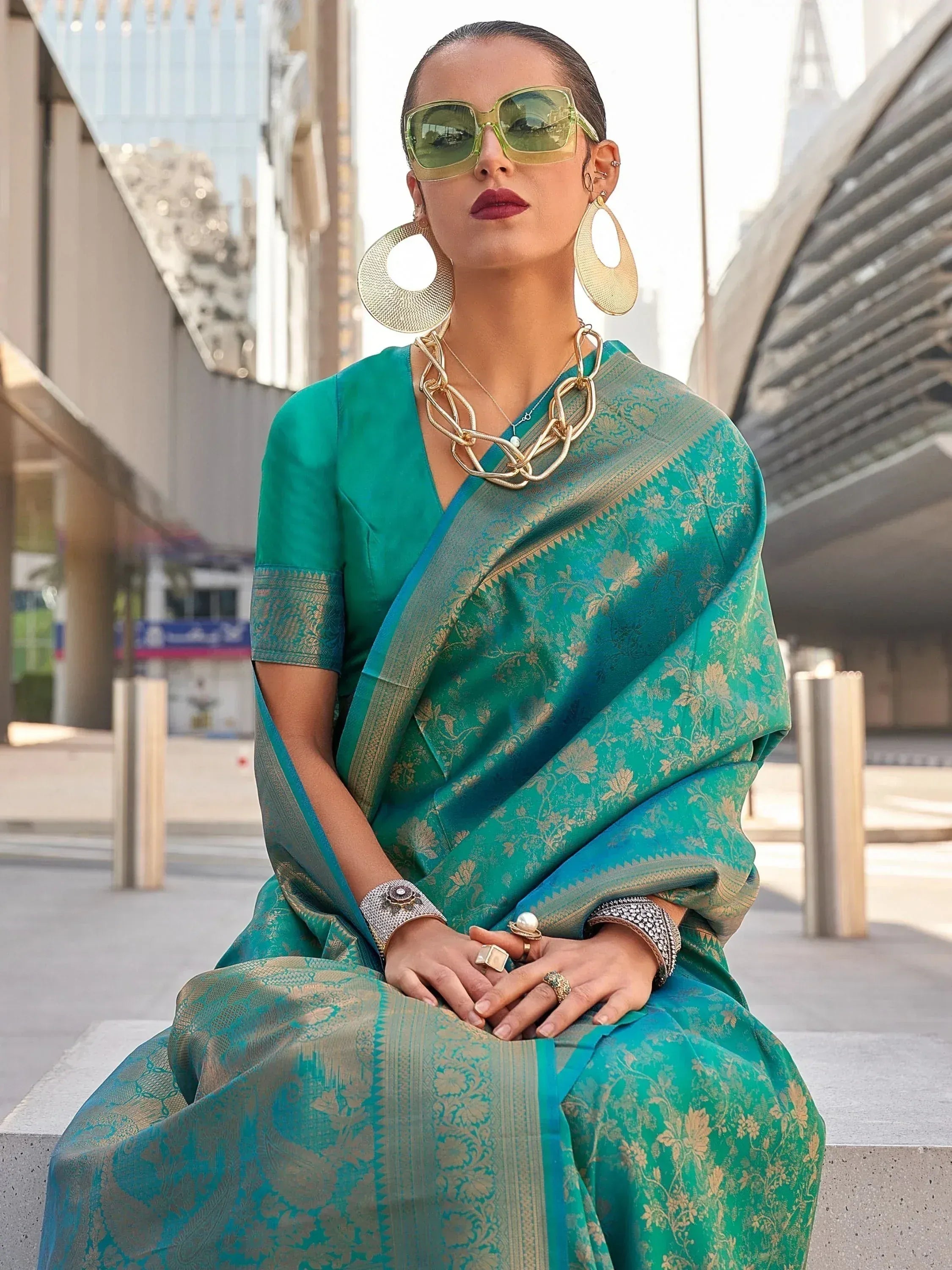 Woman in teal designer saree with gold floral print, styled with statement jewelry and sunglasses