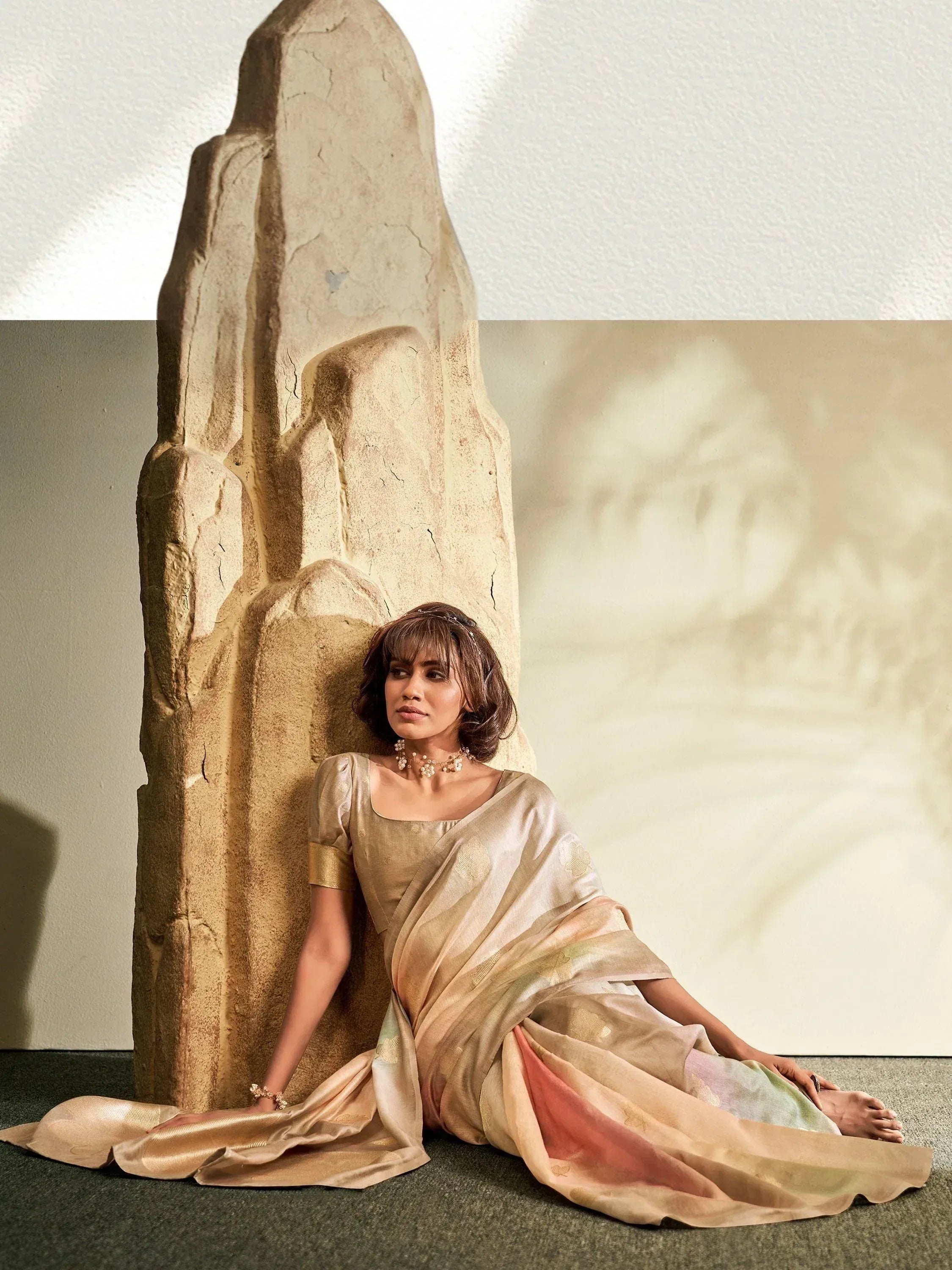 Woman in elegant gold saree with pastel ombre pallu, styled with pearl jewelry, seated beside stone sculpture