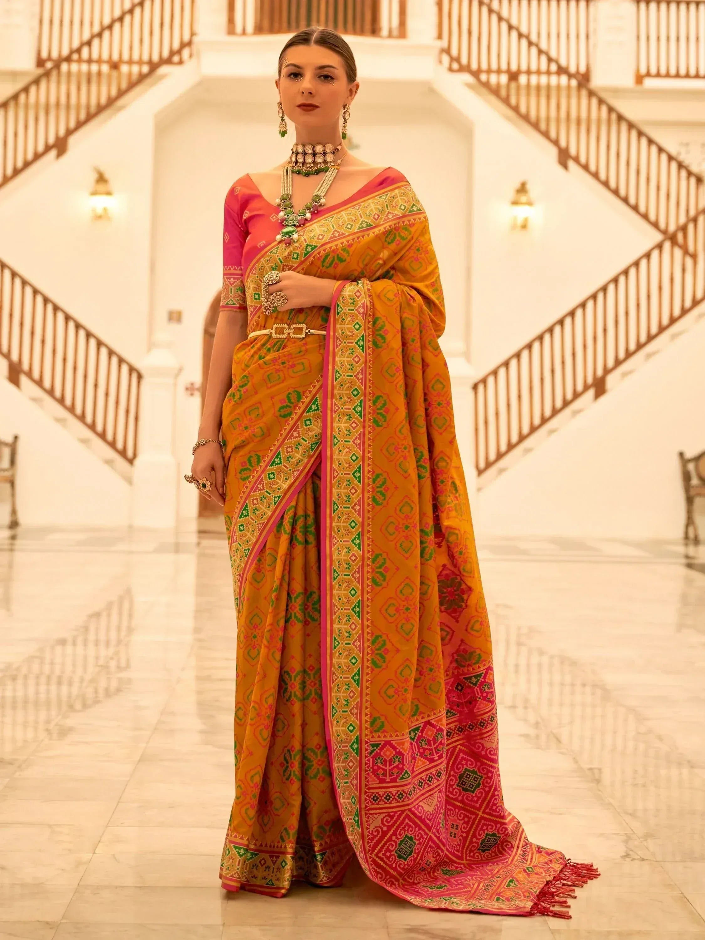Woman in vibrant yellow and pink ethnic saree with green patterns, traditional jewelry, standing in grand hall