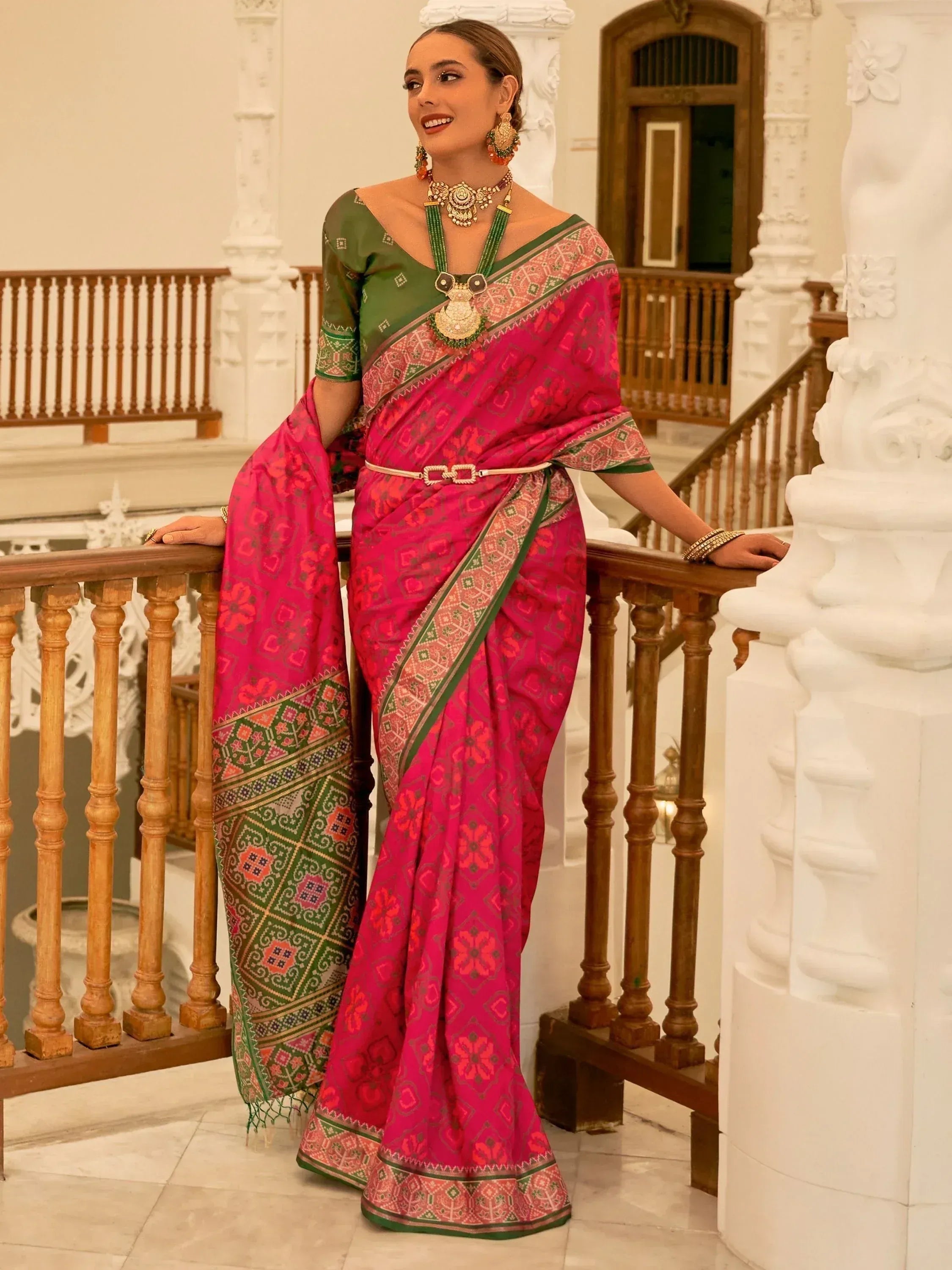 Woman in vibrant pink and green ethnic saree with intricate patterns, styled with traditional jewelry on a grand staircase.