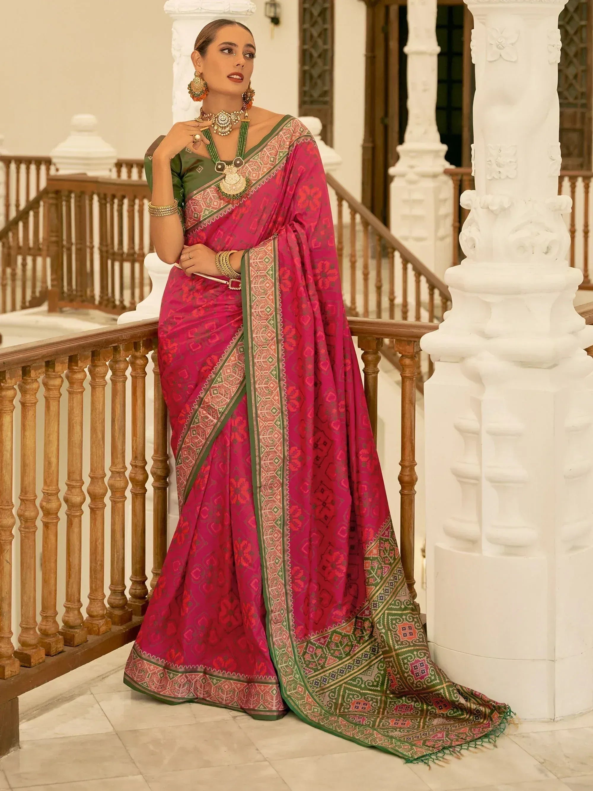 Woman in vibrant pink and green designer saree with intricate patterns, traditional jewelry, standing by ornate staircase