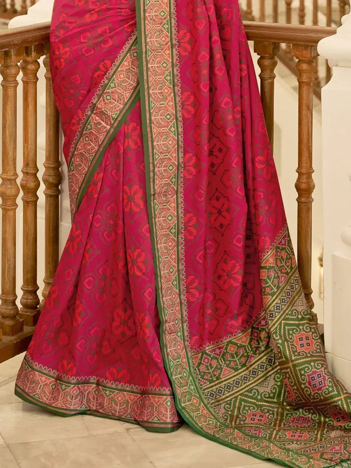Vibrant pink ethnic saree with green border and intricate geometric patterns, draped elegantly on staircase railing