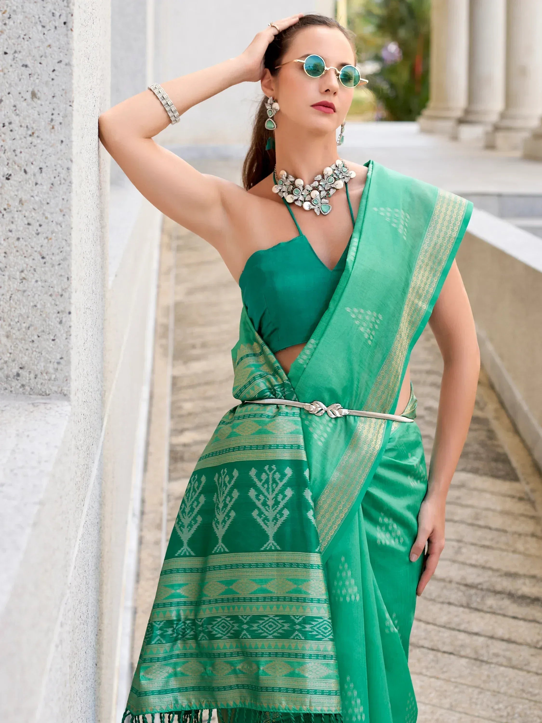 Woman in vibrant green ethnic saree with gold border, paired with statement jewelry and sunglasses, styled for modern Indian fashion.