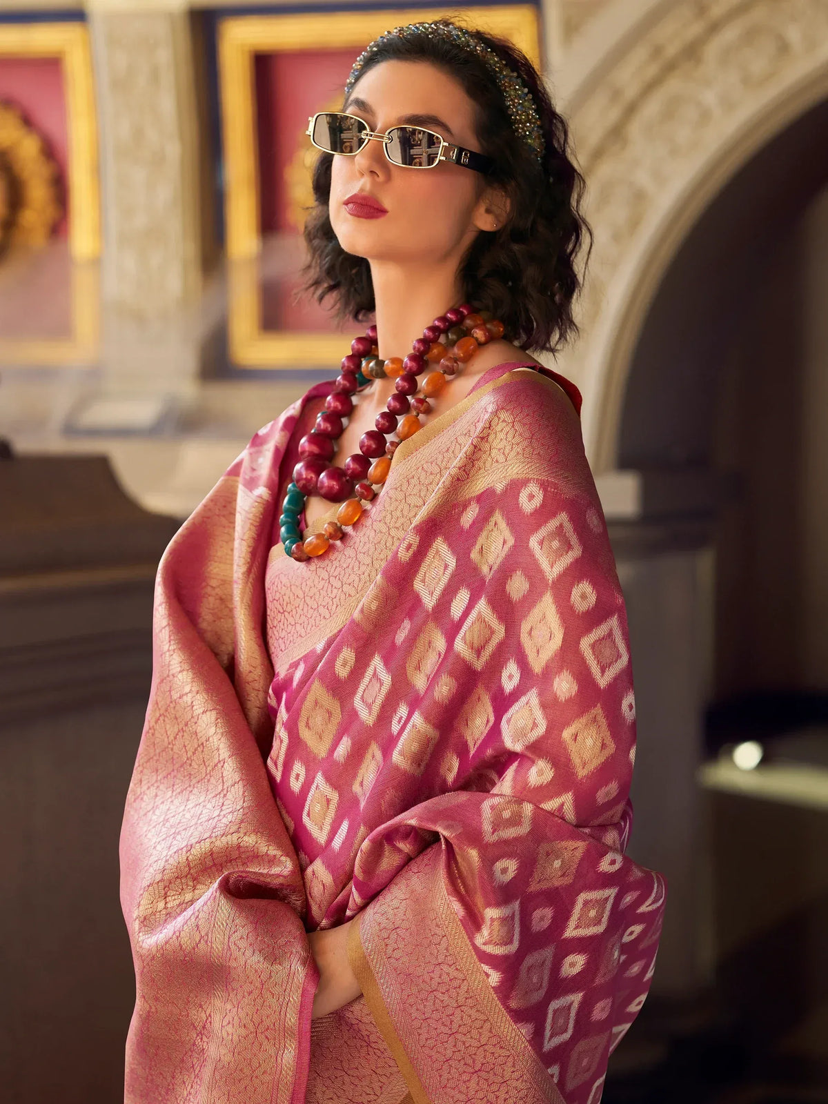 Elegant woman in pink and gold ethnic saree with geometric patterns, accessorized with beads and sunglasses.