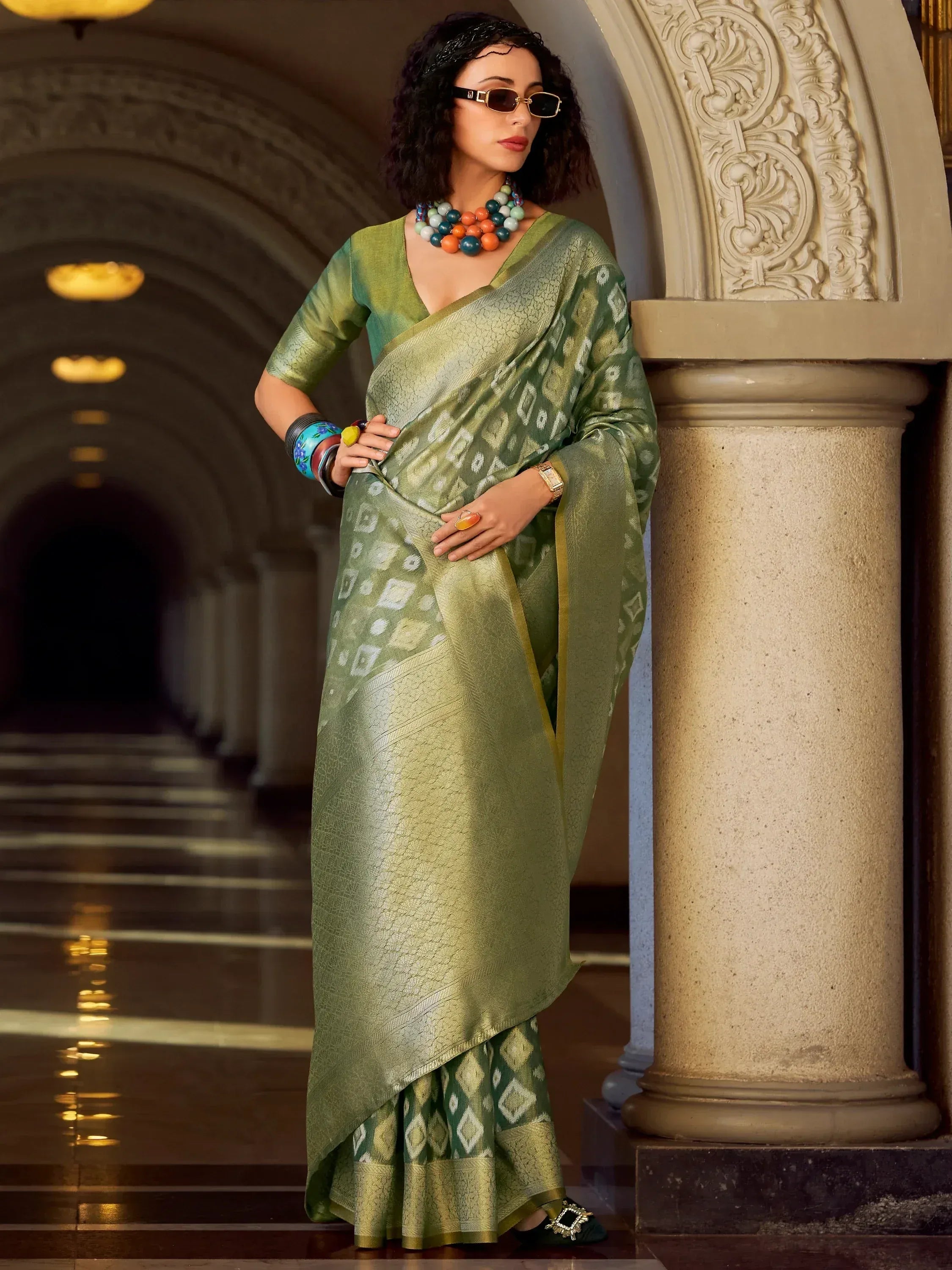 Woman in olive green designer saree with geometric print, paired with statement jewelry, in elegant hallway
