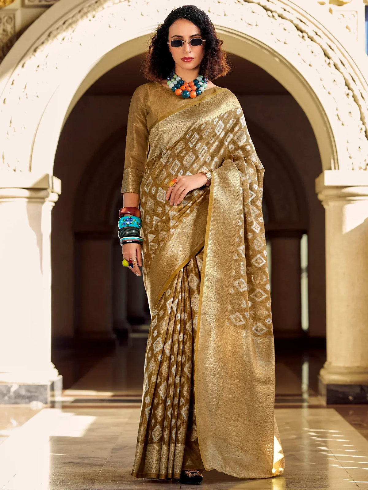 Woman in gold ethnic saree with geometric print, styled with colorful jewelry under arched corridor