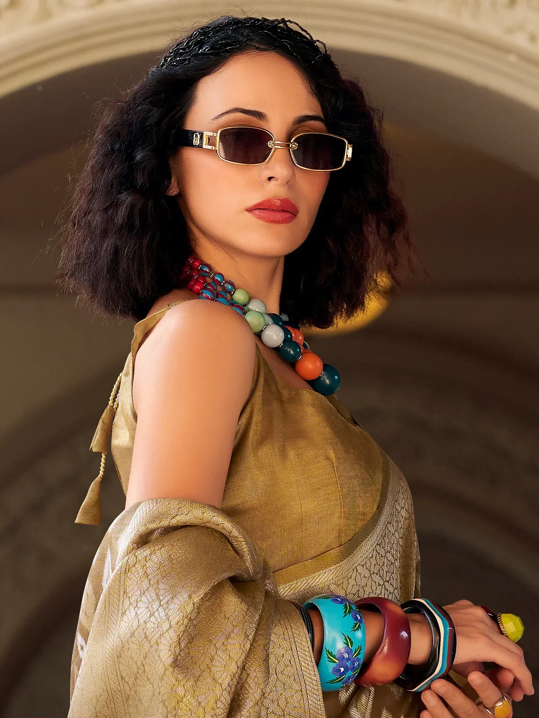 Woman in gold ethnic gown with colorful beaded necklace, bangles, and sunglasses for festive Indian wear