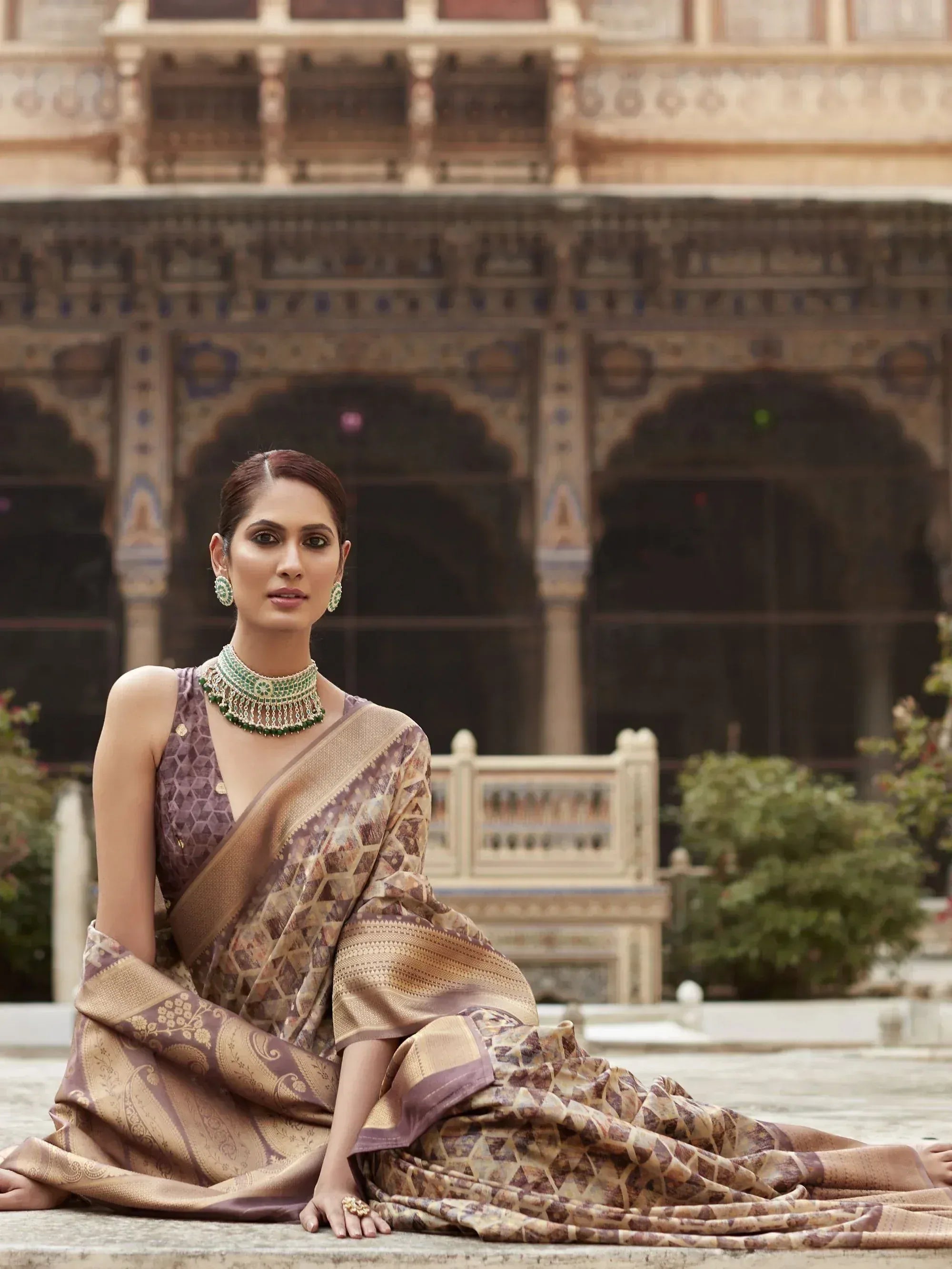 Woman in elegant Indian saree with geometric print, styled with green jewelry, seated at palace courtyard.