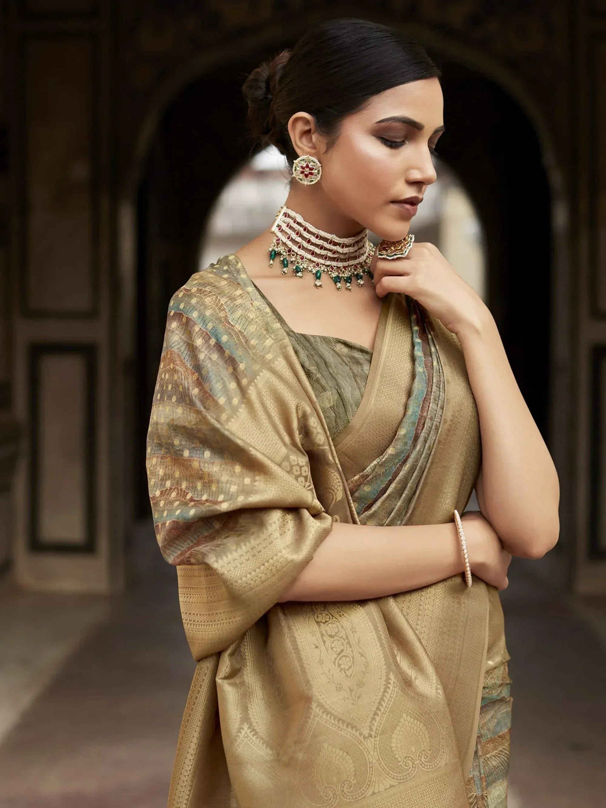 Elegant woman in gold designer saree with intricate patterns, traditional jewelry, and temple arch backdrop.
