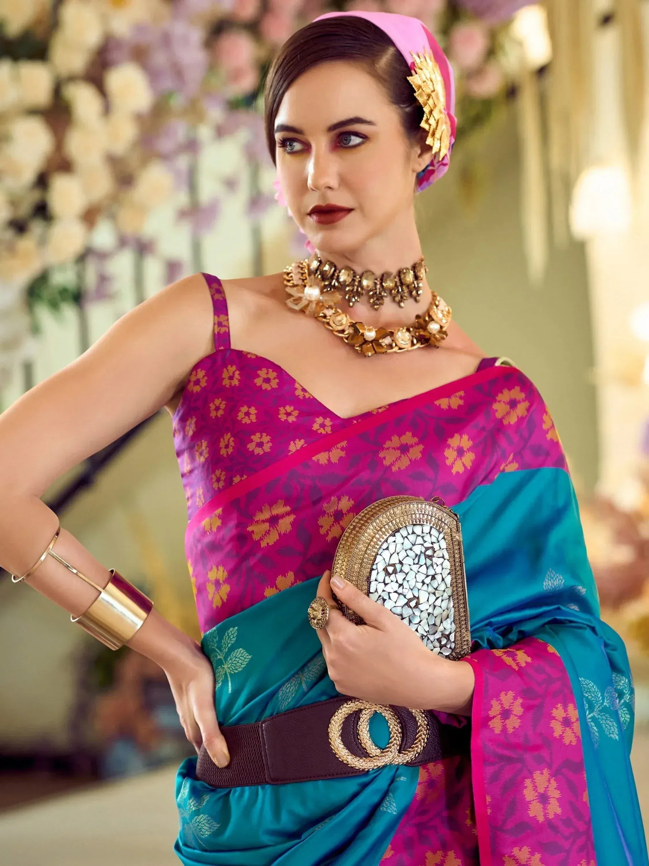 Woman in vibrant pink and teal saree with gold jewelry, floral headwrap, and clutch, styled for ethnic occasions