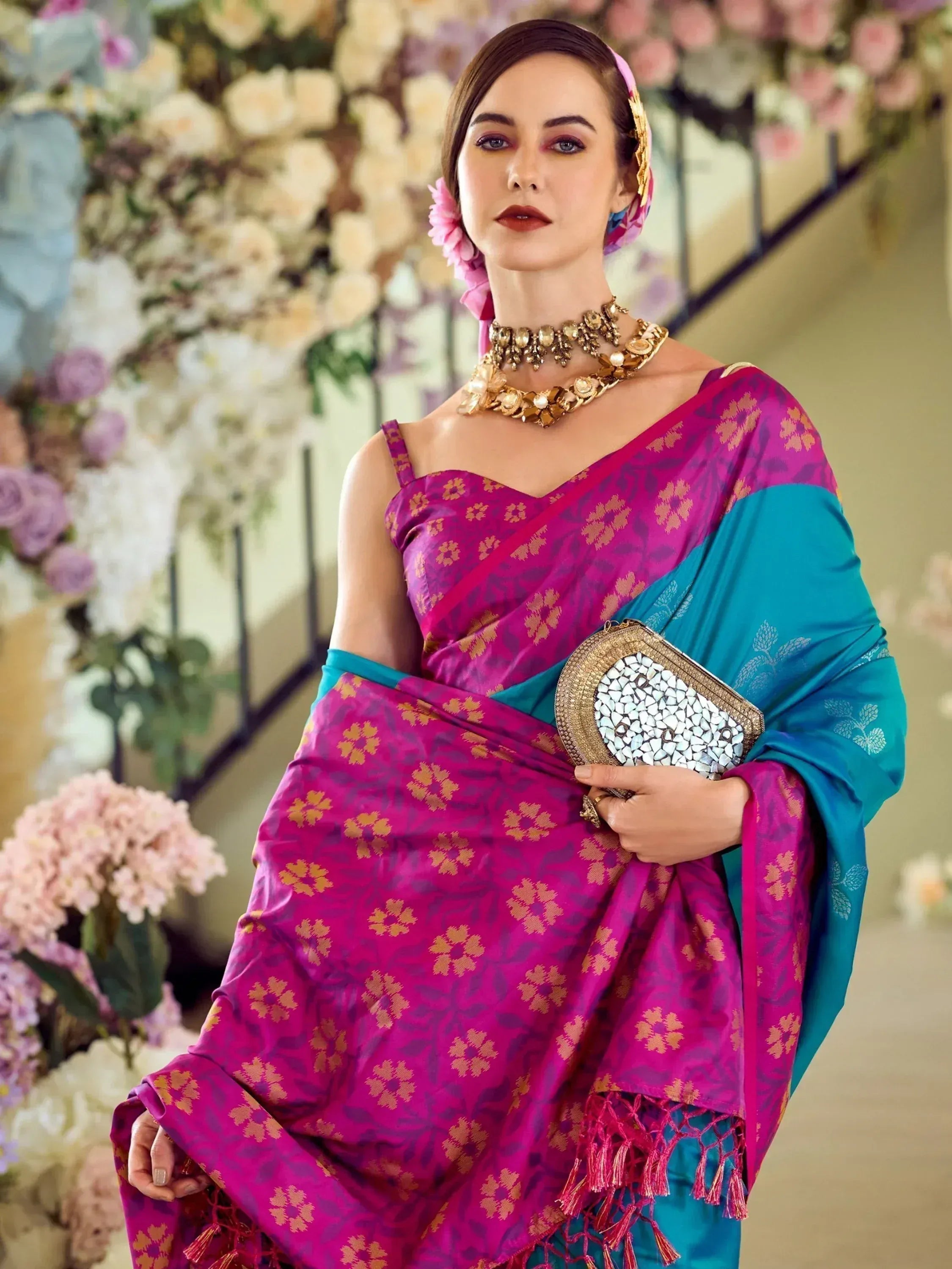 Woman in vibrant pink and teal floral saree with gold jewelry, holding clutch, floral backdrop