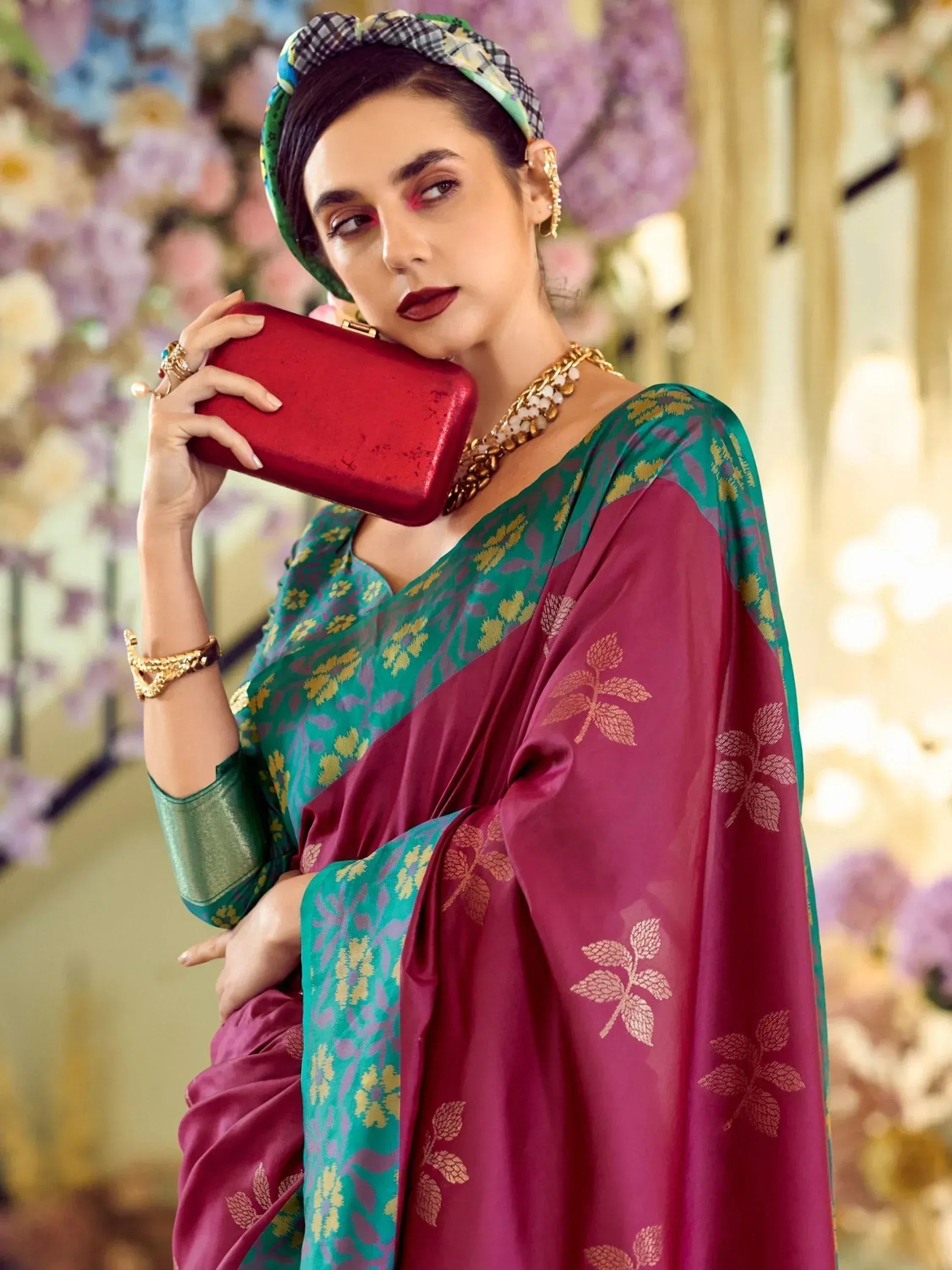 Woman in elegant green and maroon saree with gold leaf print, holding red clutch, styled with headband and gold jewelry