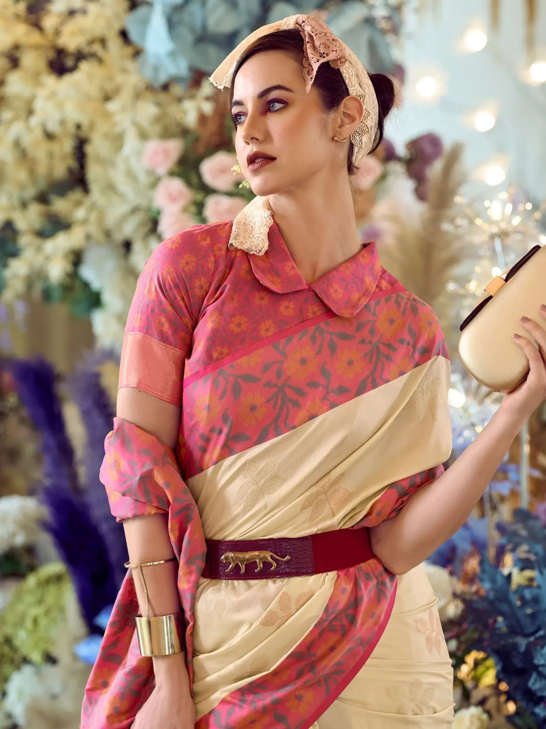 Woman in pink floral ethnic gown with lace collar, gold belt, and headscarf, holding clutch against floral backdrop