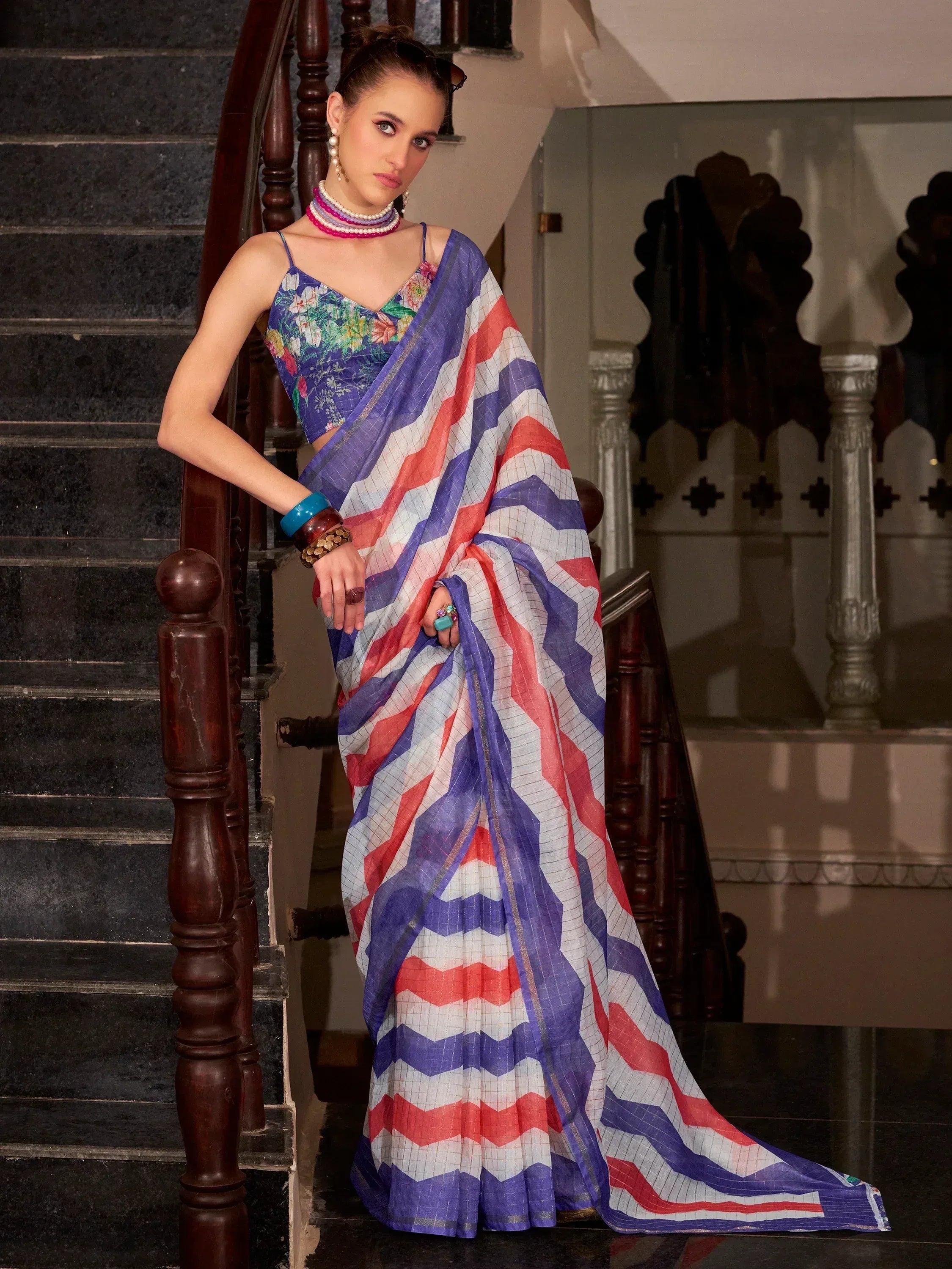Woman in vibrant ethnic saree with zigzag patterns, floral blouse, and traditional jewelry on staircase