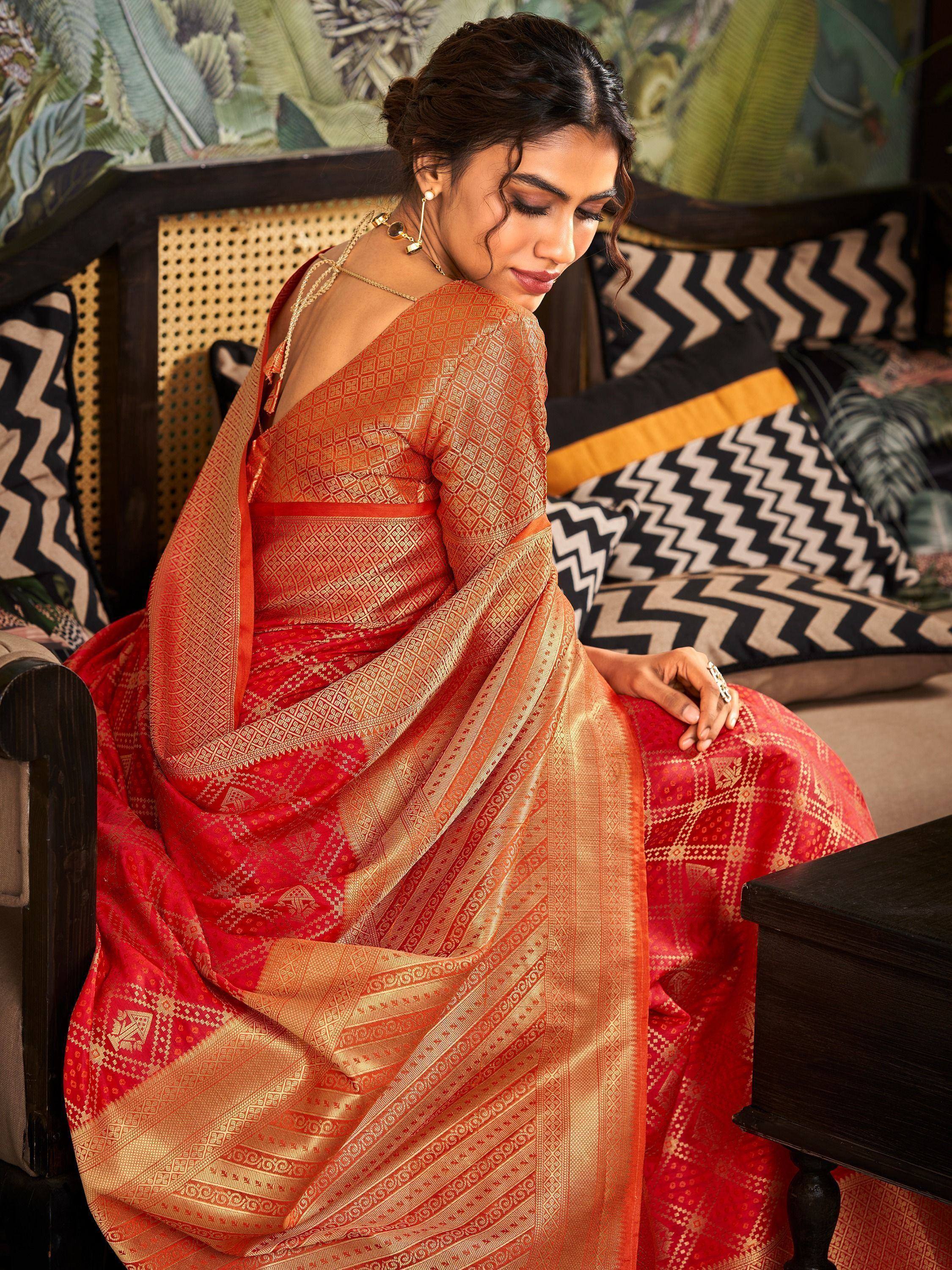 Woman in red and gold ethnic saree with intricate patterns, seated on patterned sofa