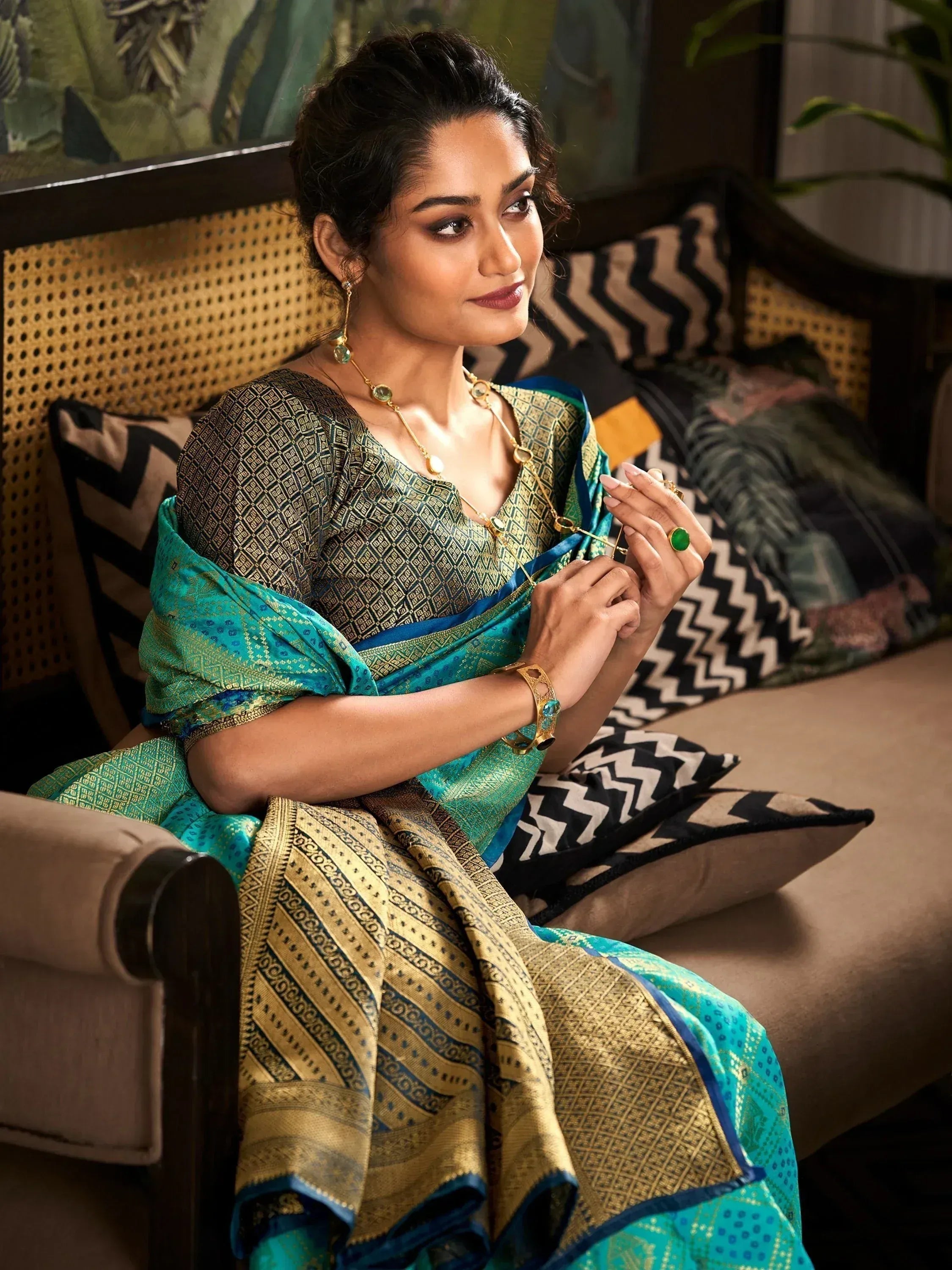 Woman in elegant teal and gold designer saree with traditional jewelry, seated on a stylish sofa