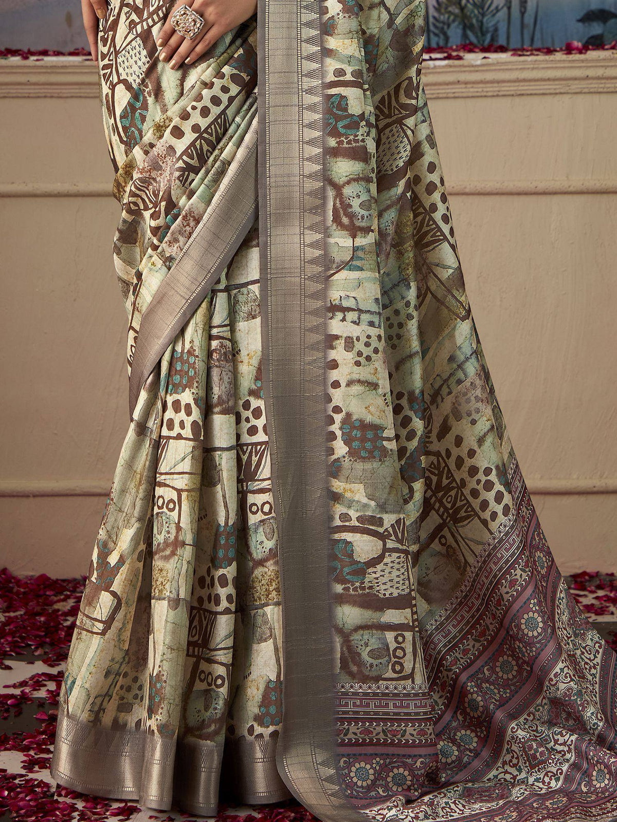Elegant printed saree with abstract brown and teal motifs, metallic border, and floral pallu, styled with rose petals.