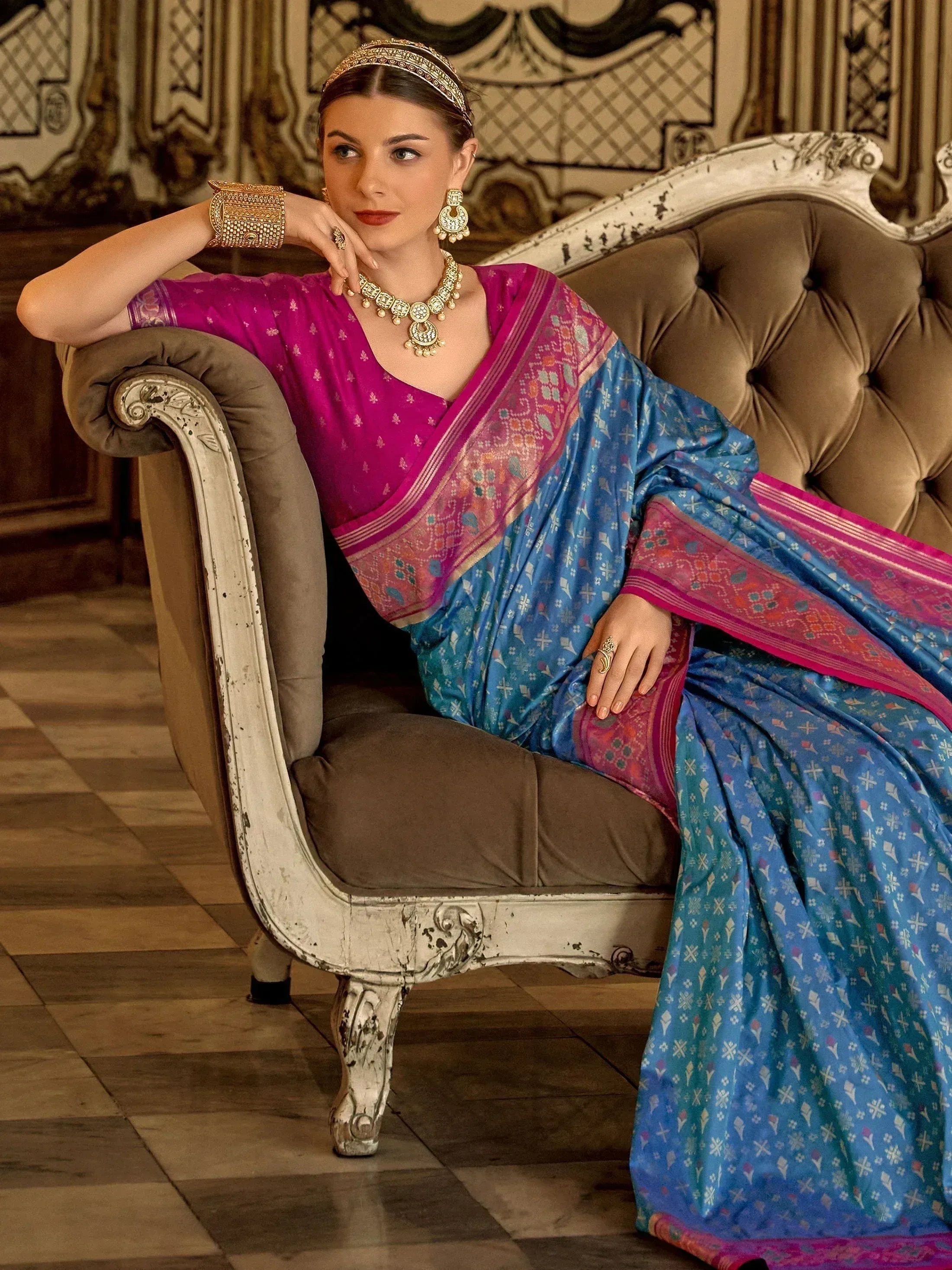 Woman in vibrant blue and pink printed saree with gold jewelry, seated on vintage sofa