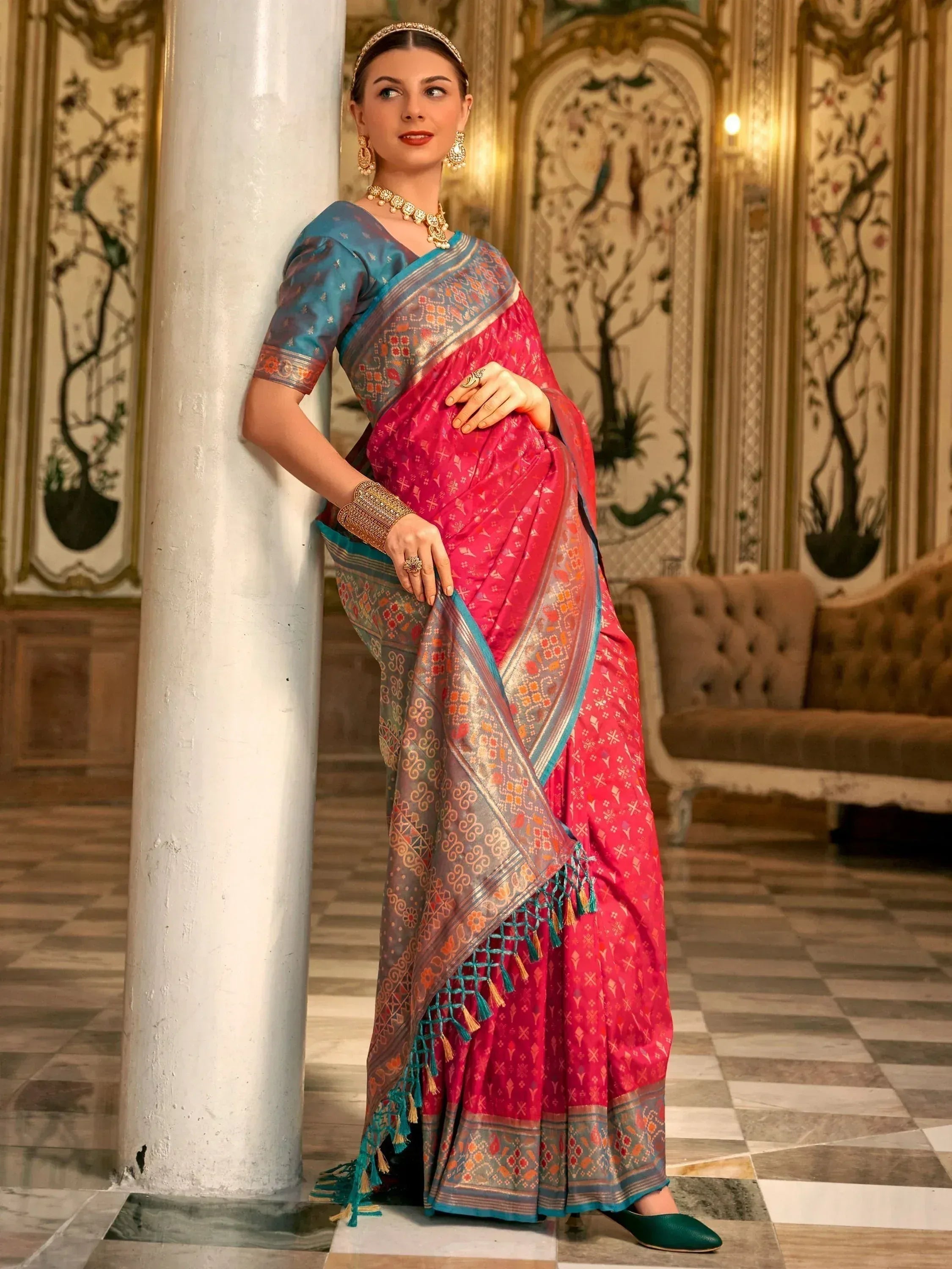 Woman in vibrant pink and blue ethnic saree with intricate patterns, traditional jewelry, leaning against pillar