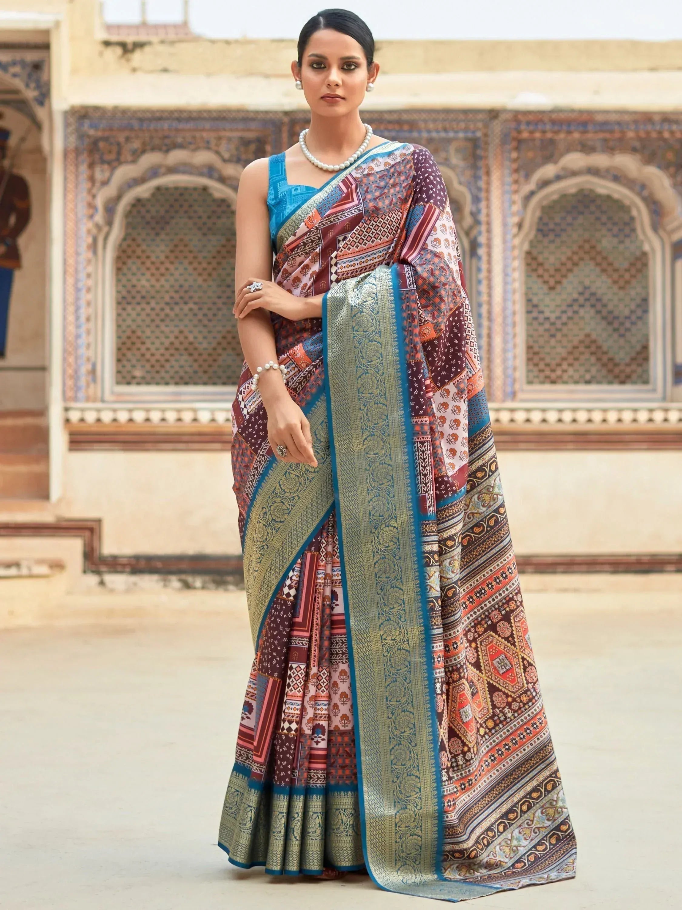 Woman in colorful printed saree with blue blouse, ethnic jewelry, standing before ornate palace backdrop