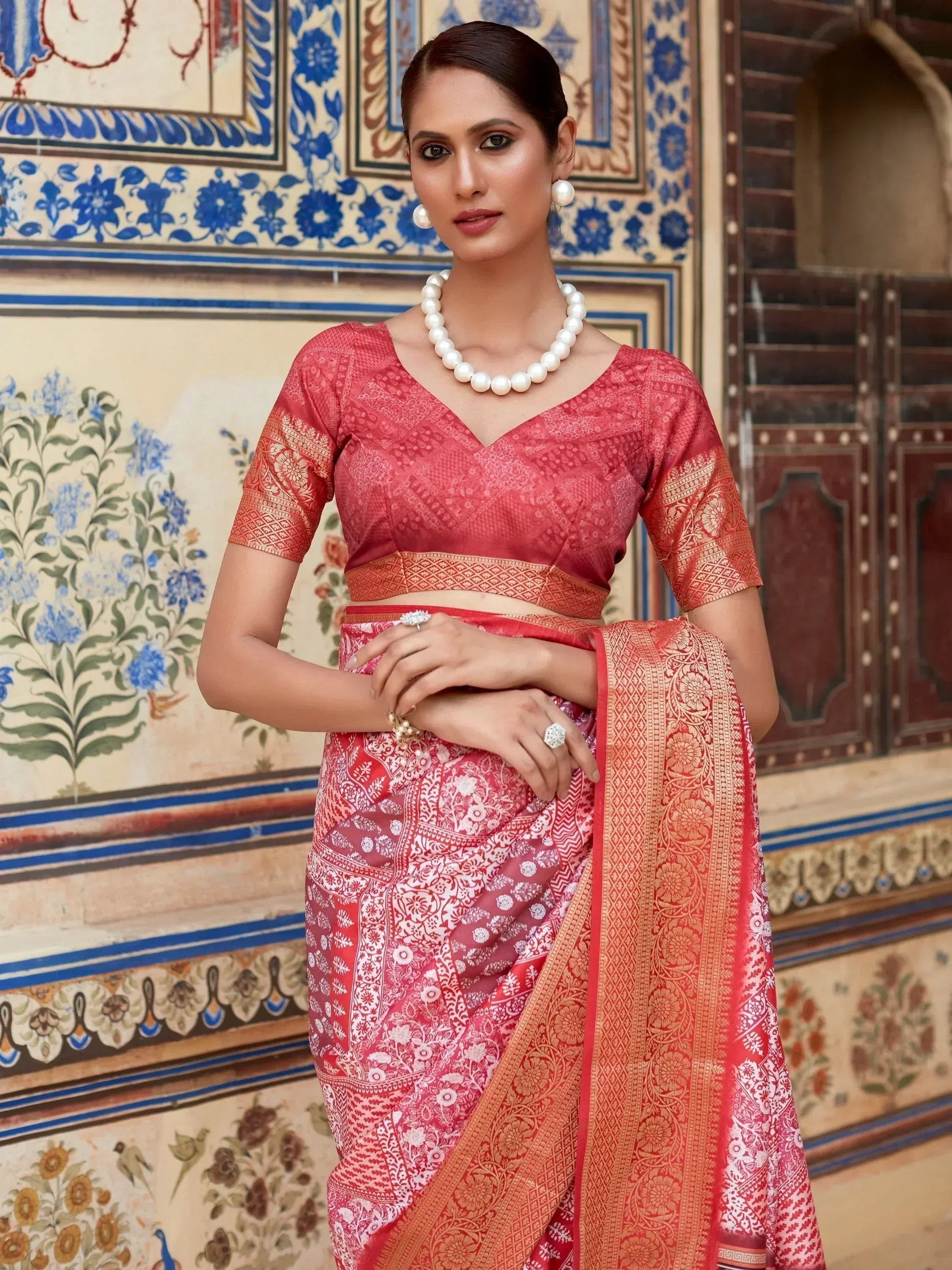 Woman in red and gold printed saree with V-neck blouse, pearl jewelry, against ornate blue floral wall