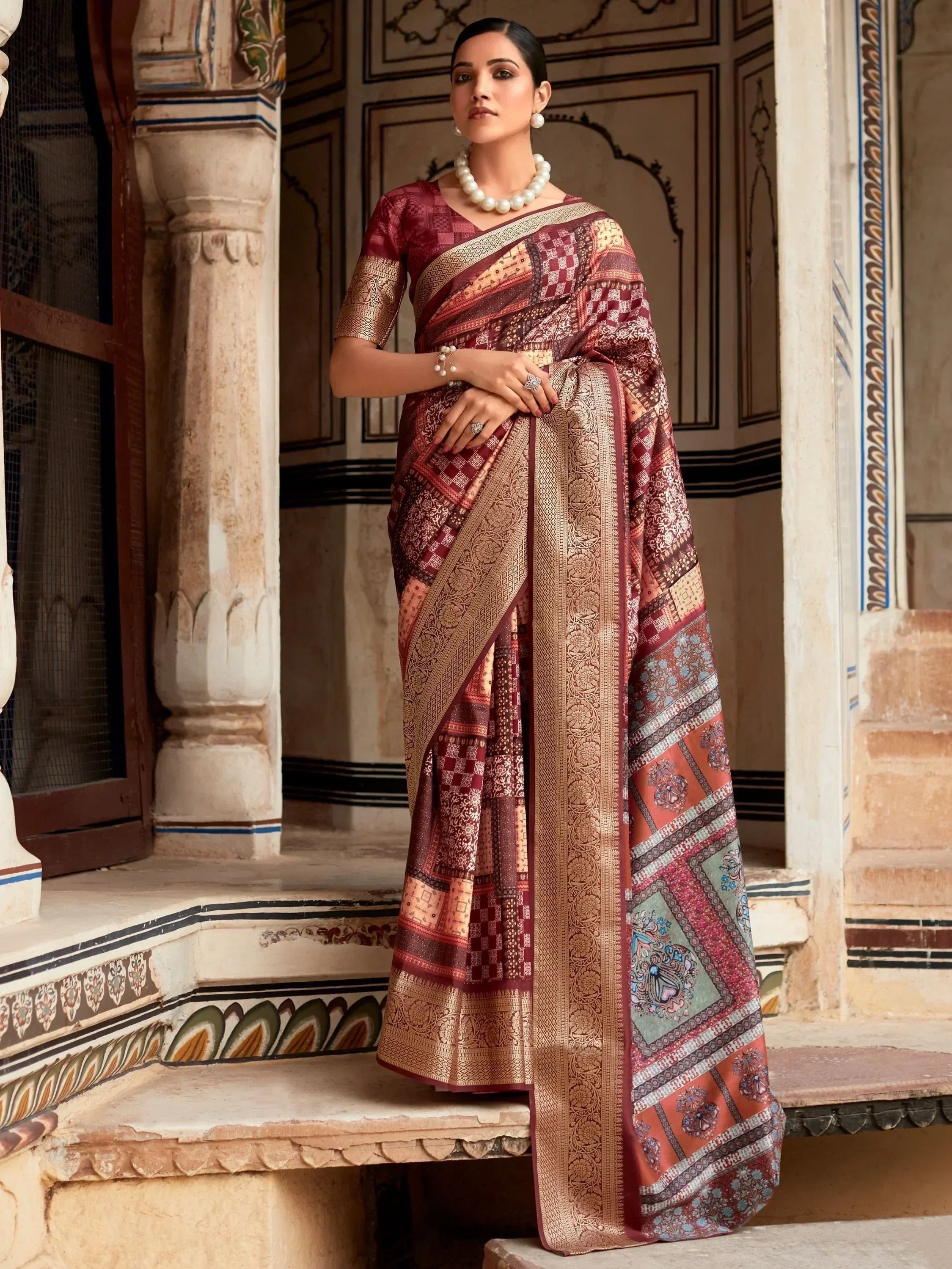 Woman in maroon and gold designer saree with intricate patterns, styled with pearl jewelry at heritage Indian palace