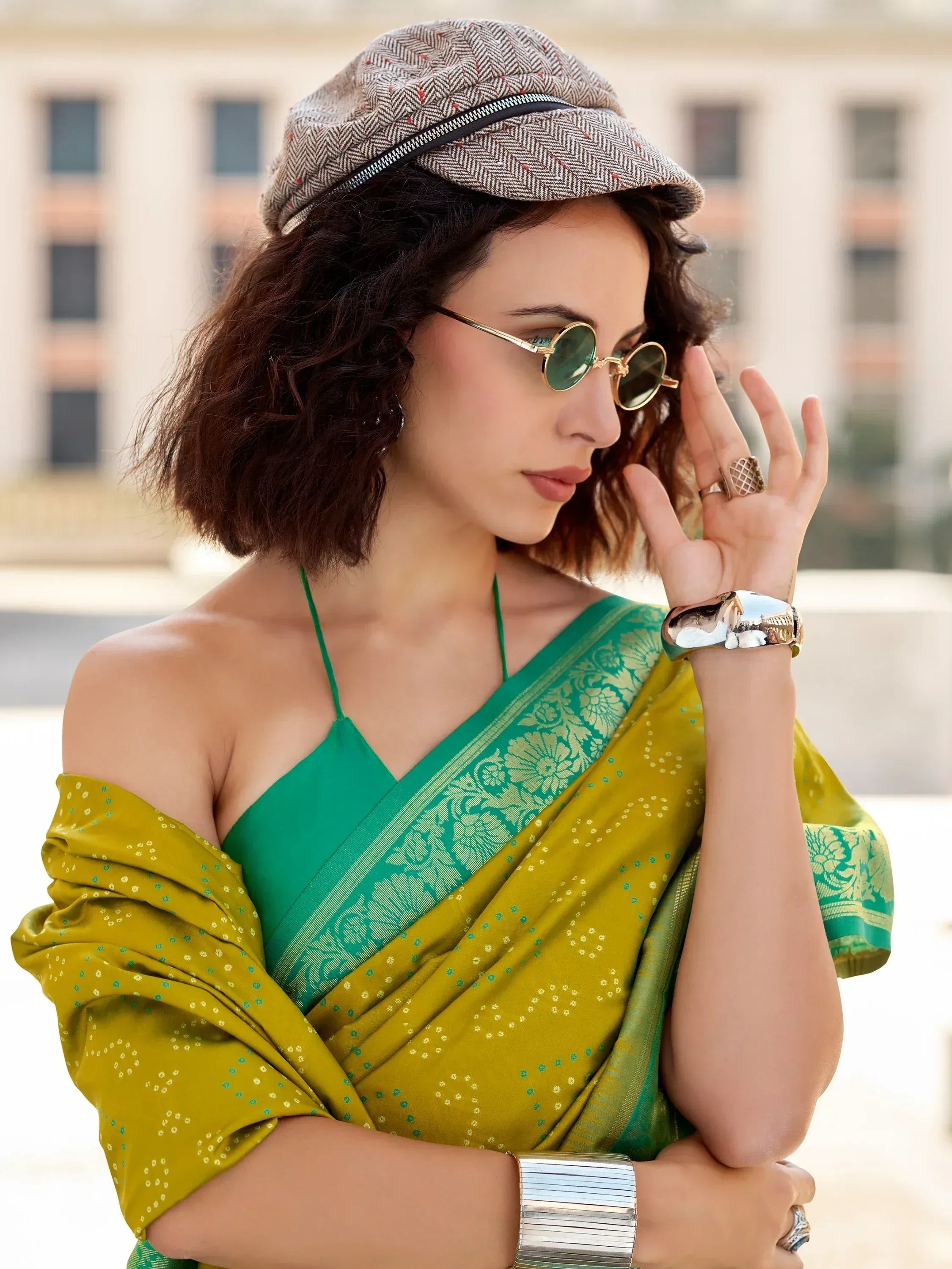 Stylish woman in green and yellow saree with cap, sunglasses, and ethnic jewelry for modern Indian wear.