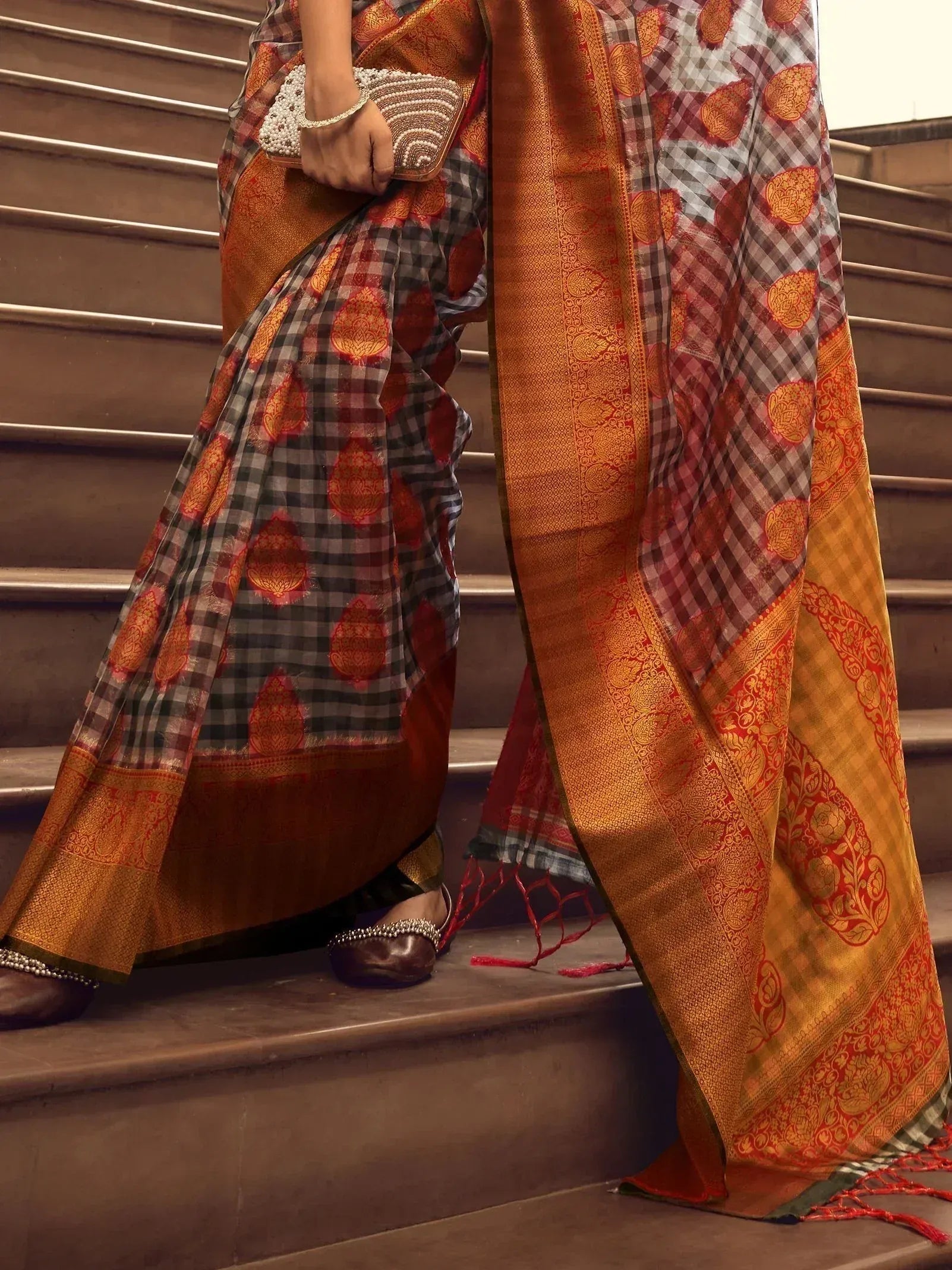 Woman in elegant ethnic saree with checkered and floral motifs, gold border, holding clutch on stairs