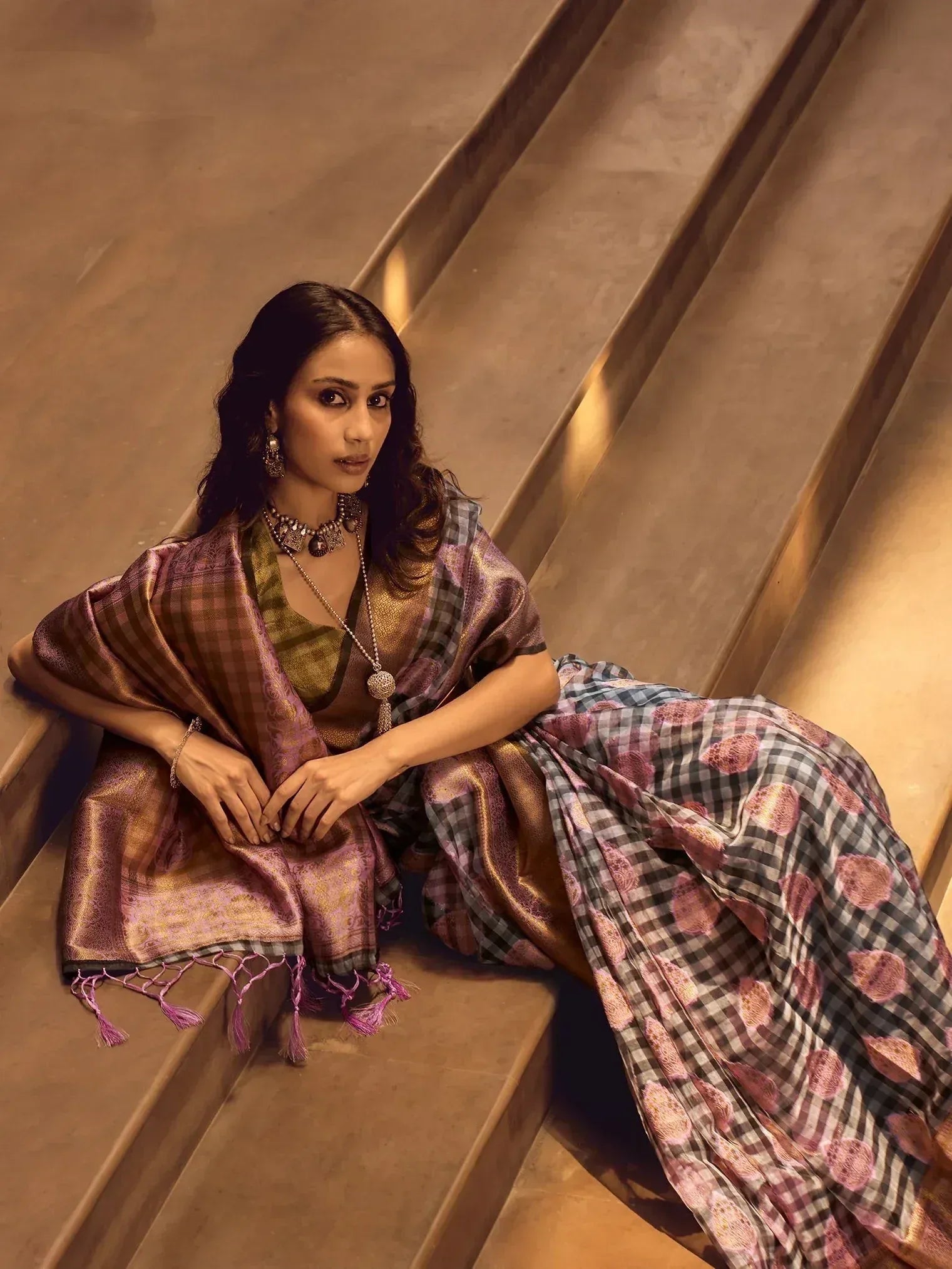 Woman in elegant Indian saree with checkered and floral print, styled with traditional jewelry, sitting on stairs