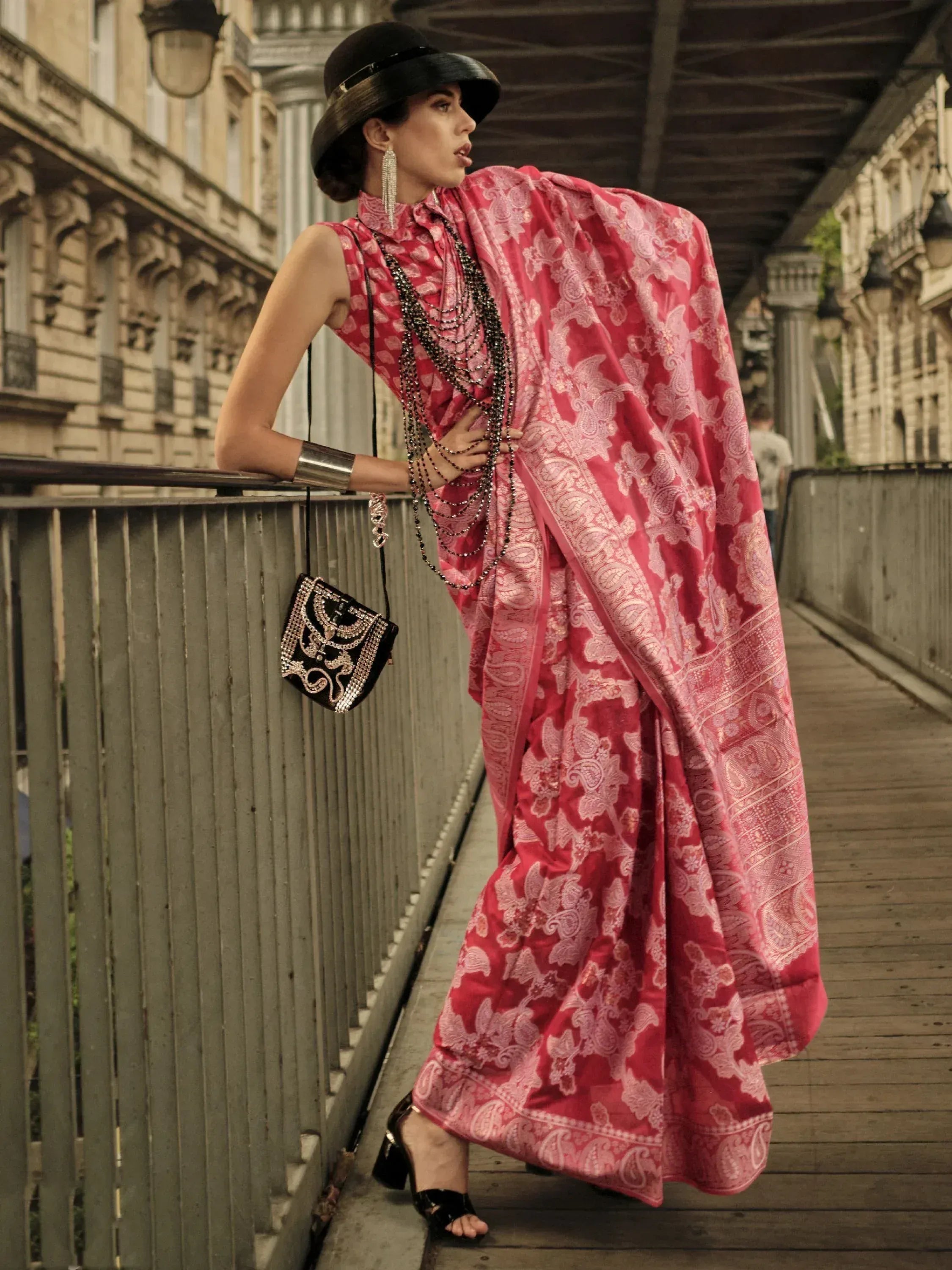 Woman in pink floral designer saree with black hat and embellished bag, urban bridge setting