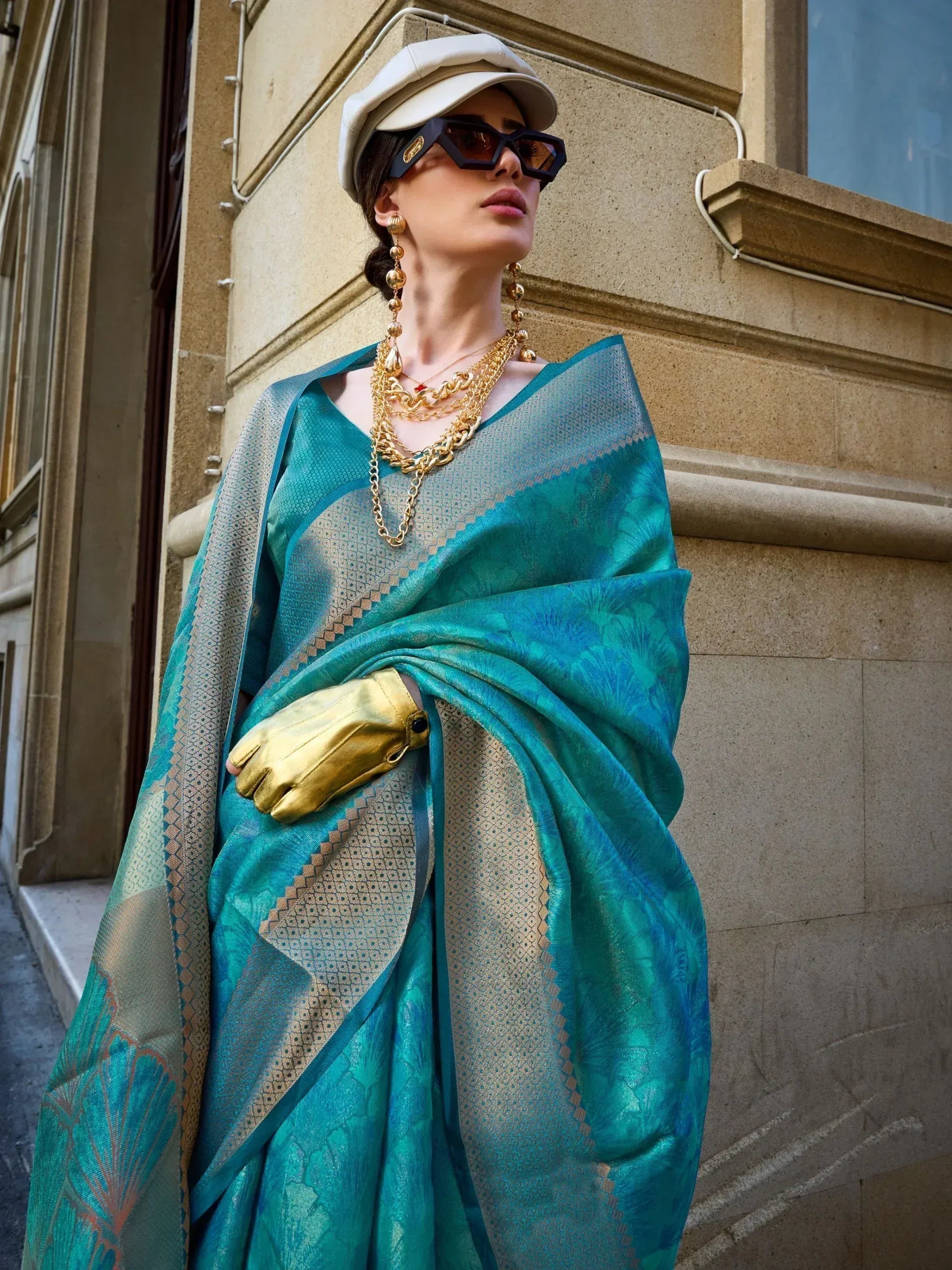 Stylish woman in teal designer saree with gold jewelry, cap, and sunglasses, showcasing ethnic fashion.