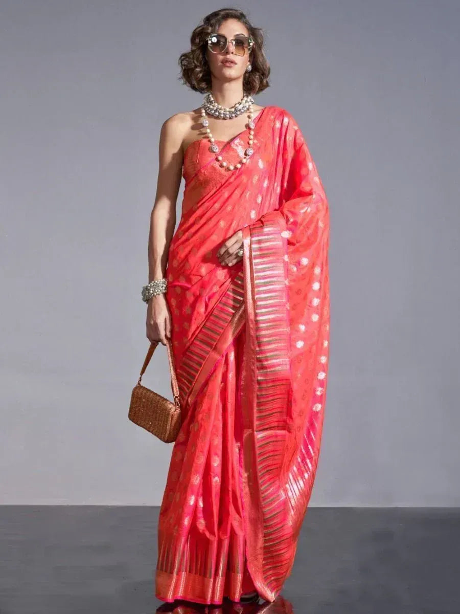 Woman in vibrant coral saree with gold motifs, pearl jewelry, and woven clutch, styled for ethnic occasions