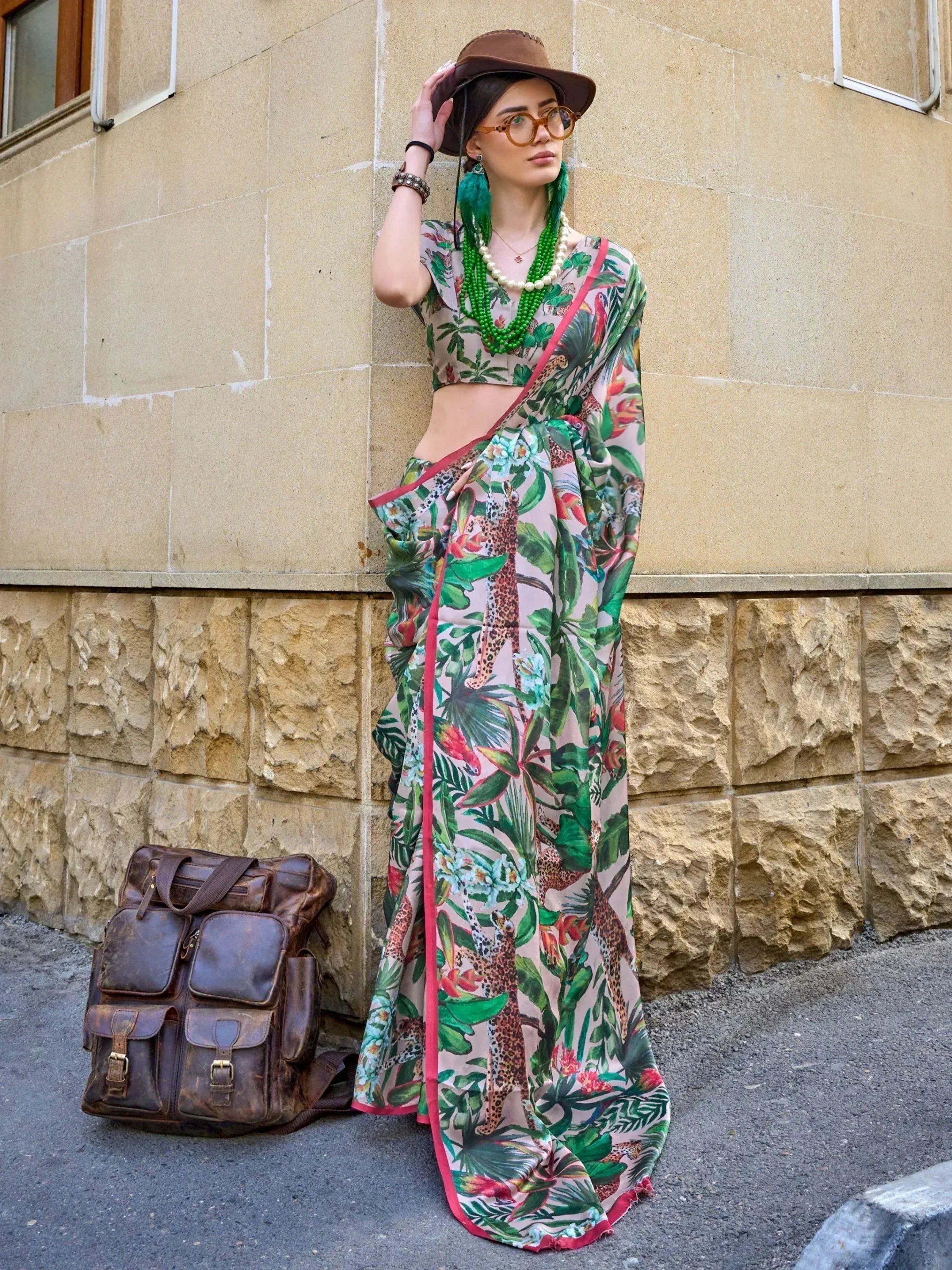 Woman in tropical print saree with leopard and foliage design, styled with hat and green beads, standing by stone wall.