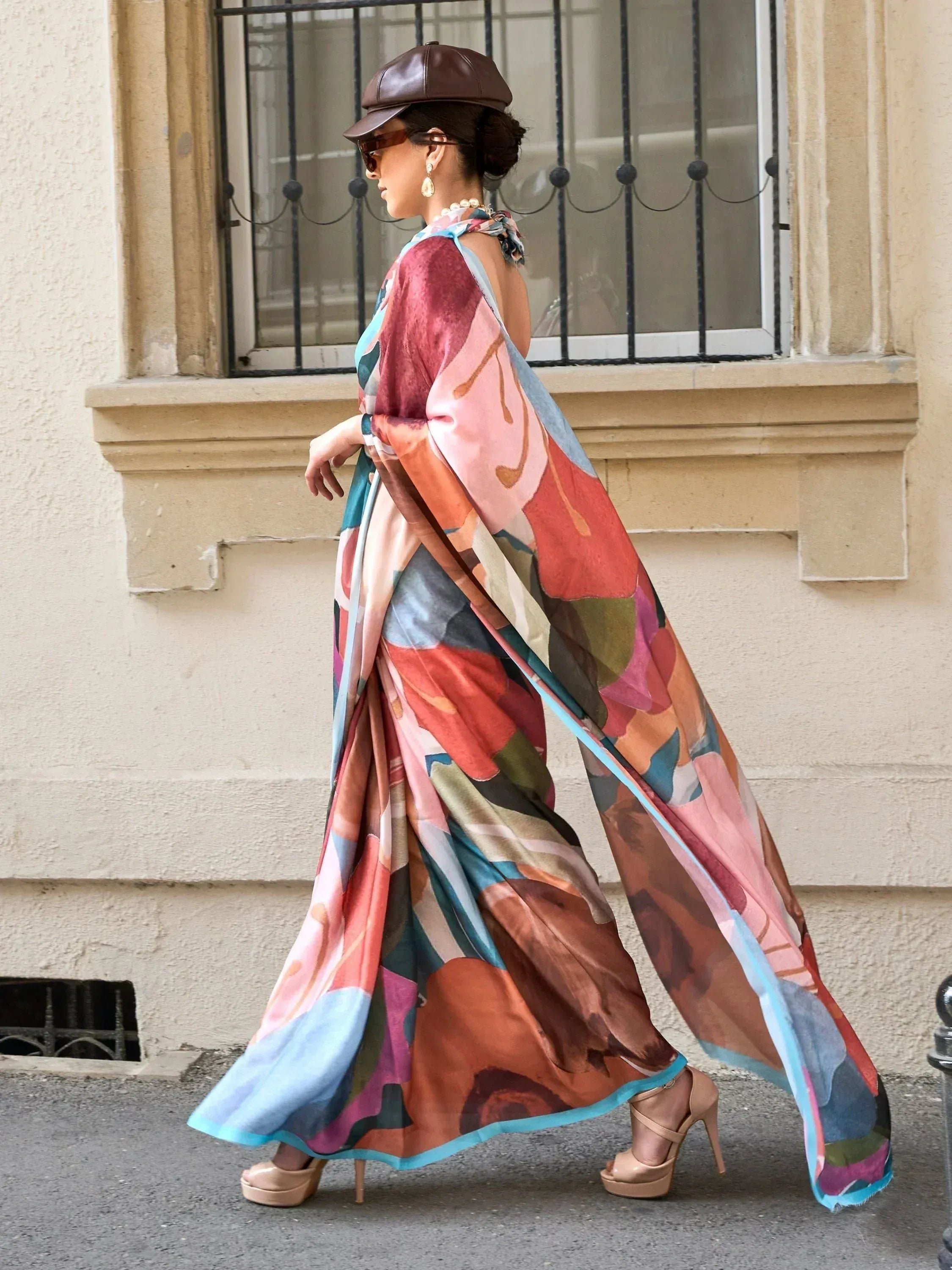 Woman in colorful abstract print ethnic gown with cape sleeves, brown cap, and heels walking outdoors