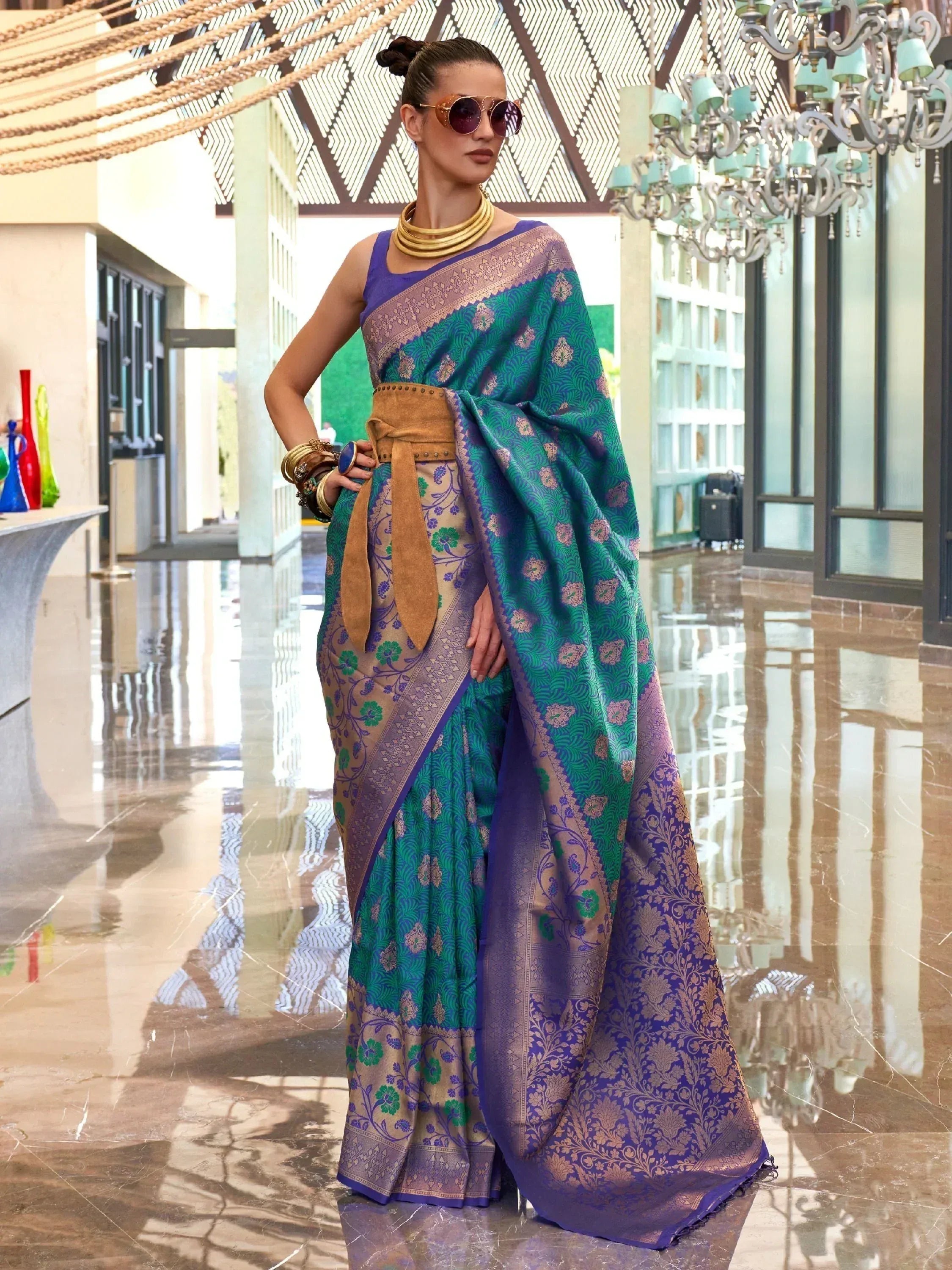 Elegant woman in designer saree with purple and green floral print, gold border, styled with belt and statement jewelry.