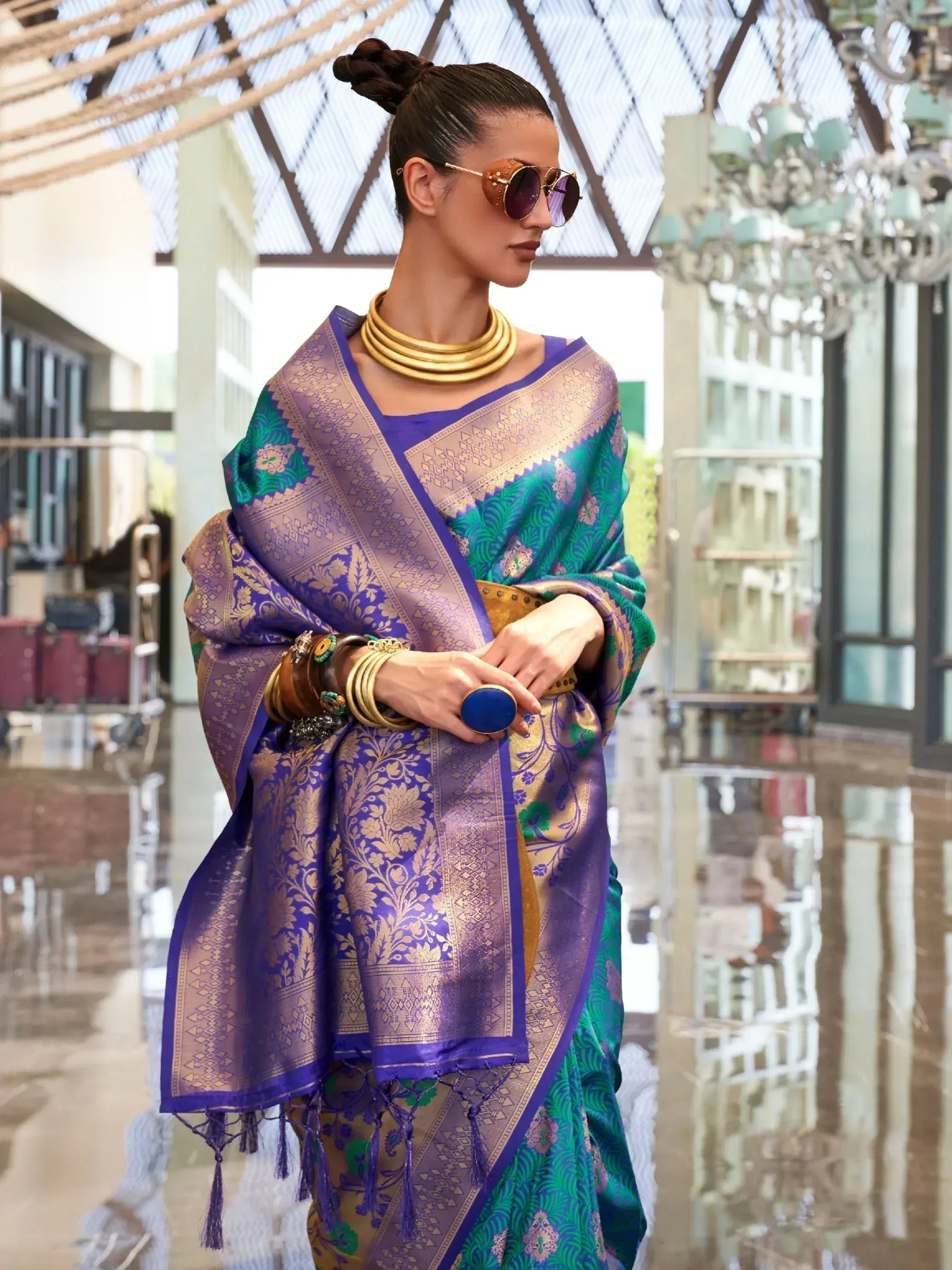 Woman in purple and green designer saree with gold embroidery, ethnic jewelry, and sunglasses