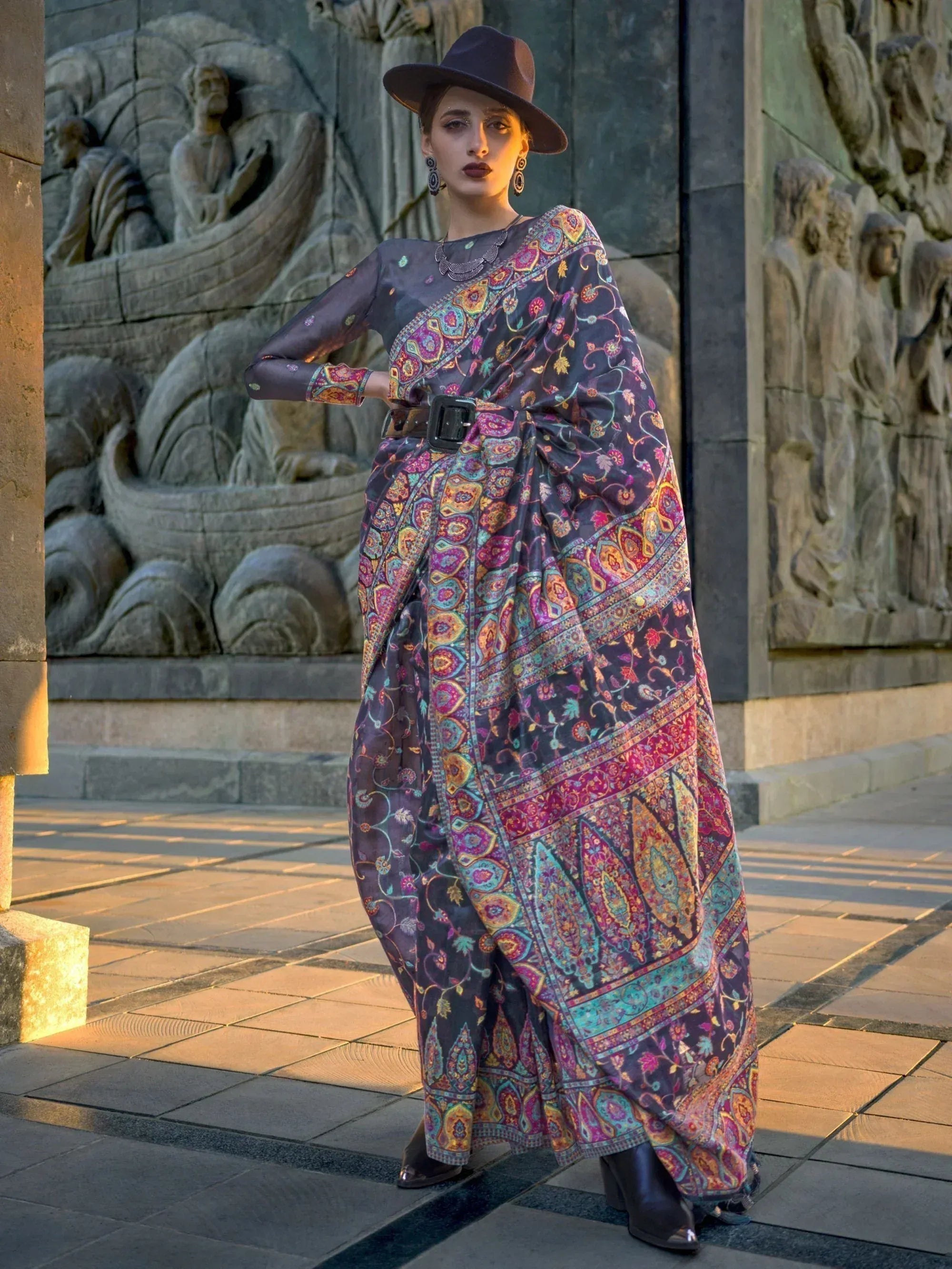 Woman in colorful printed ethnic gown with belt, hat, and boots, standing near stone carvings